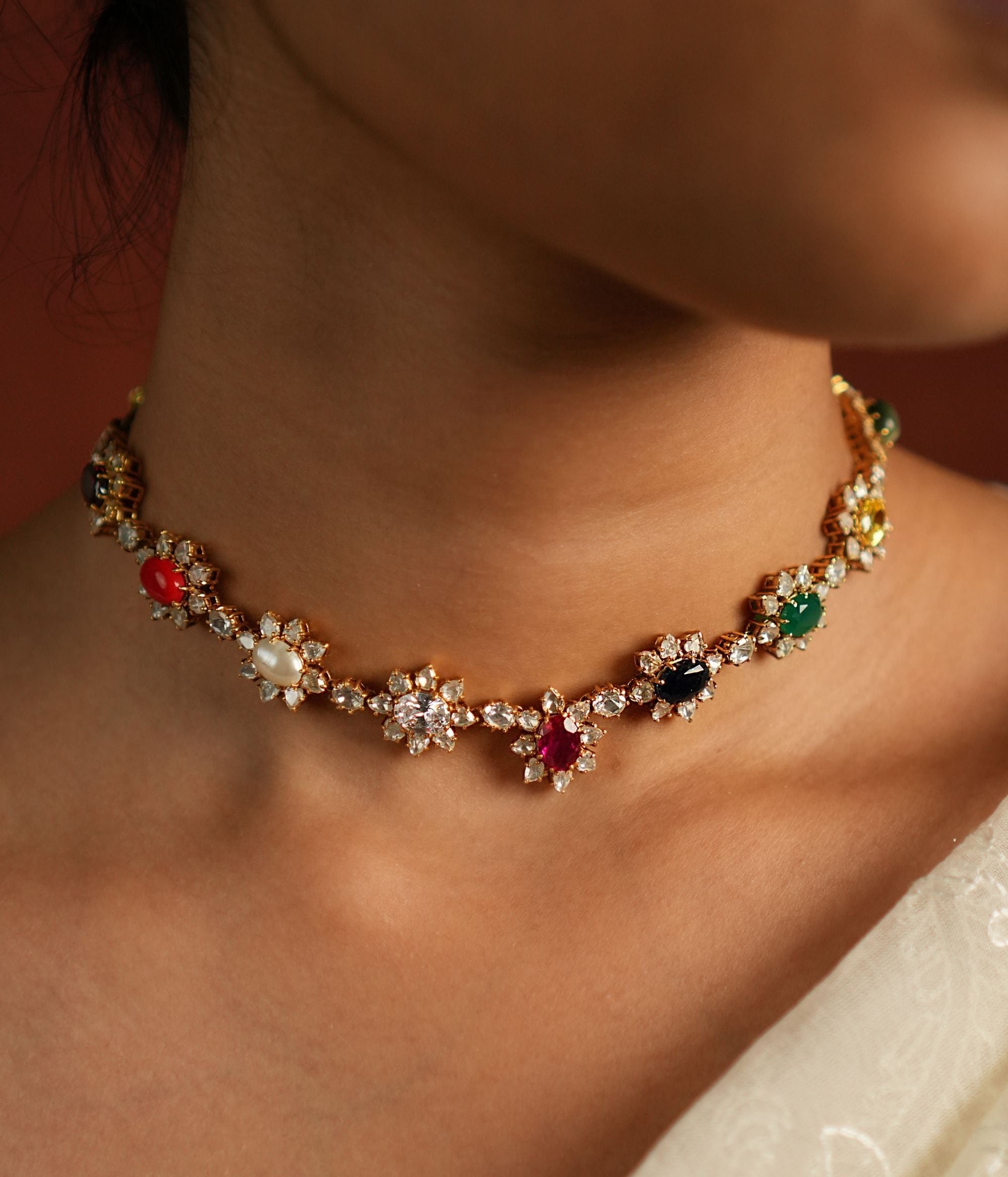 A sophisticated gold-plated silver choker featuring a delicate chain adorned with exquisite Navratna stones arranged in a charming floral design. Each vibrant gemstone is beautifully encased by shimmering CZ stones, adding a touch of elegance and brilliance. This timeless piece seamlessly blends tradition with modern sophistication, making it a perfect accessory for any occasion.