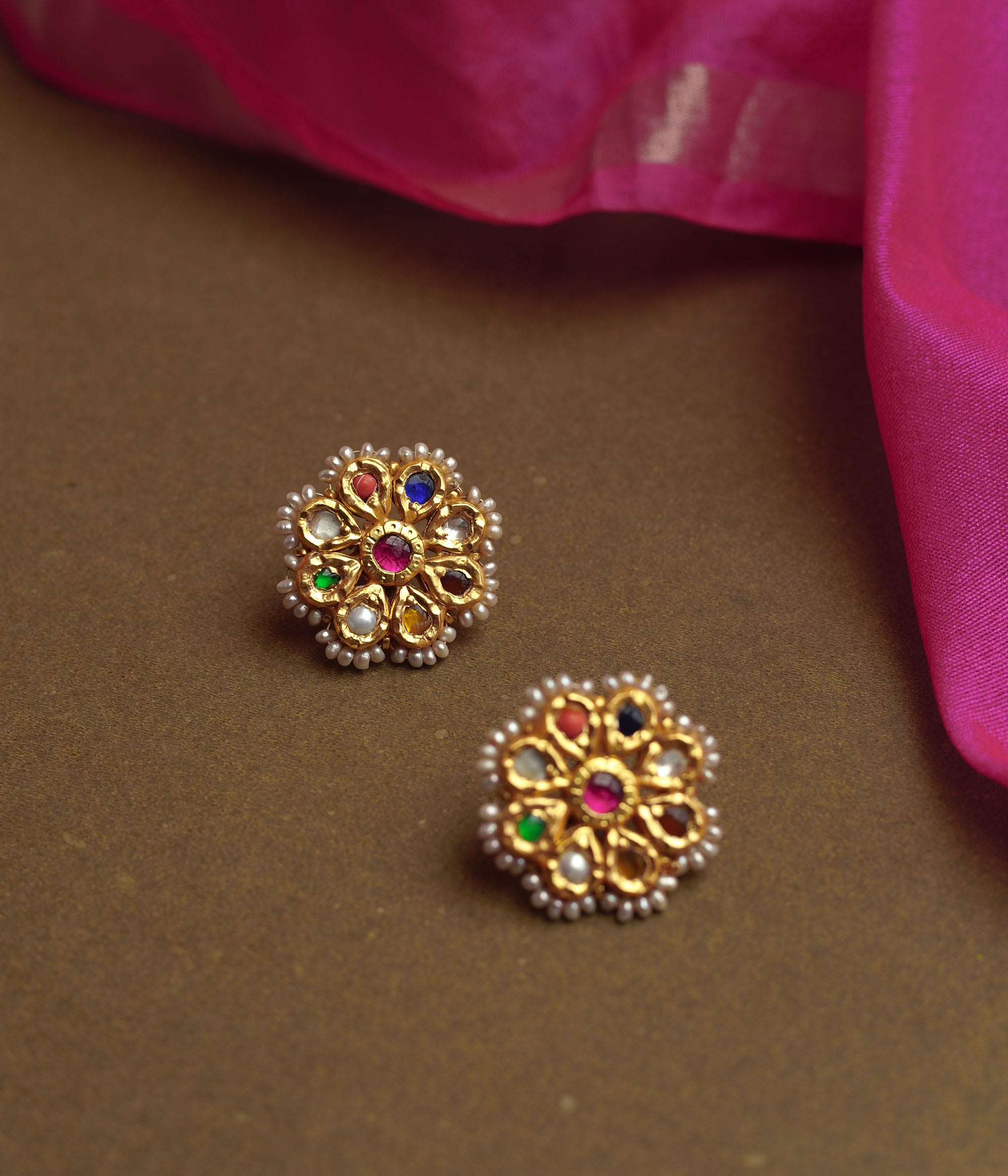 Close-up of two ornate gold stud earrings. Each features a central red gem surrounded by colorful Navratna stones and an outer border of small pearls, resting on a brown surface.