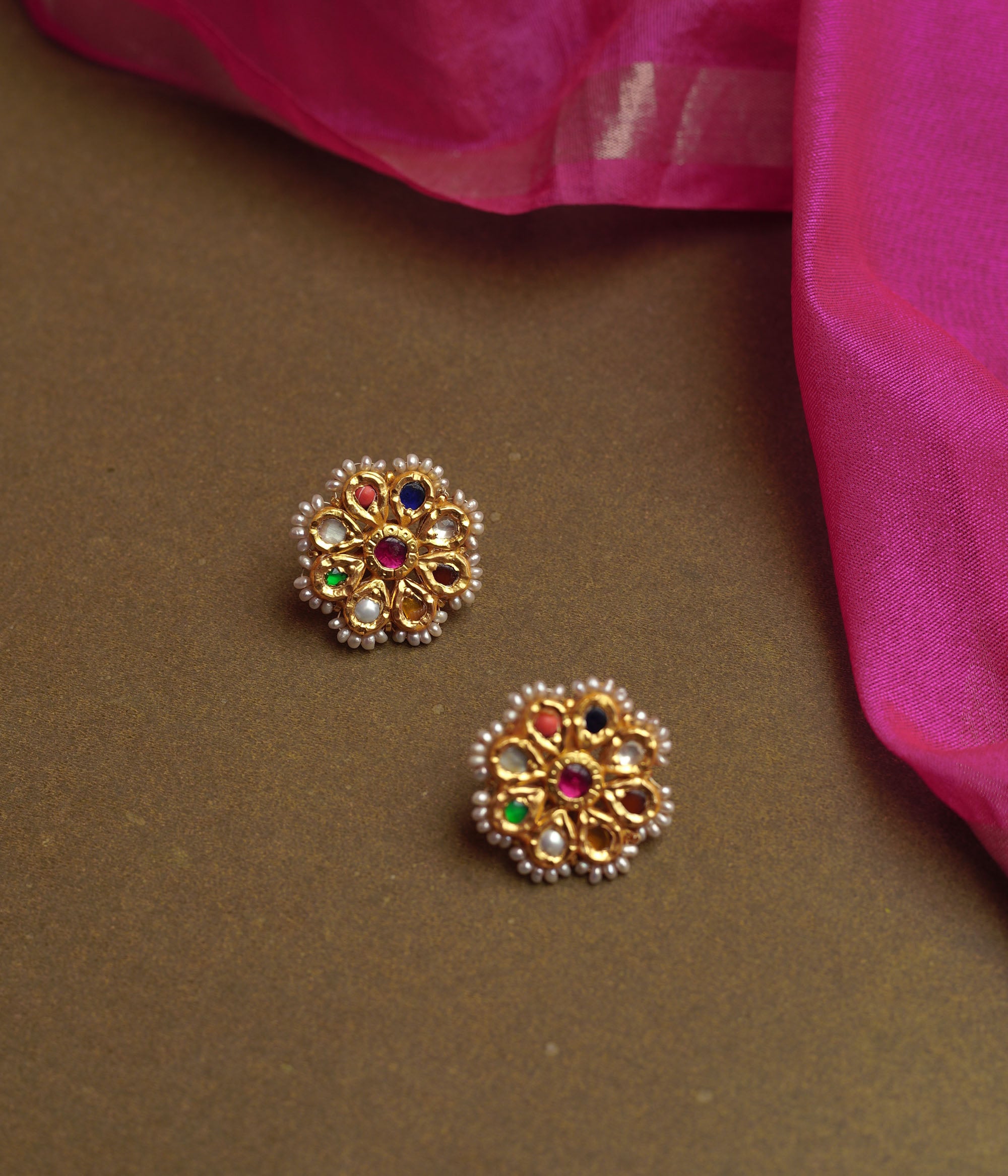 Close-up of two ornate gold stud earrings. Each features a central red gem surrounded by colorful Navratna stones and an outer border of small pearls, resting on a brown surface.