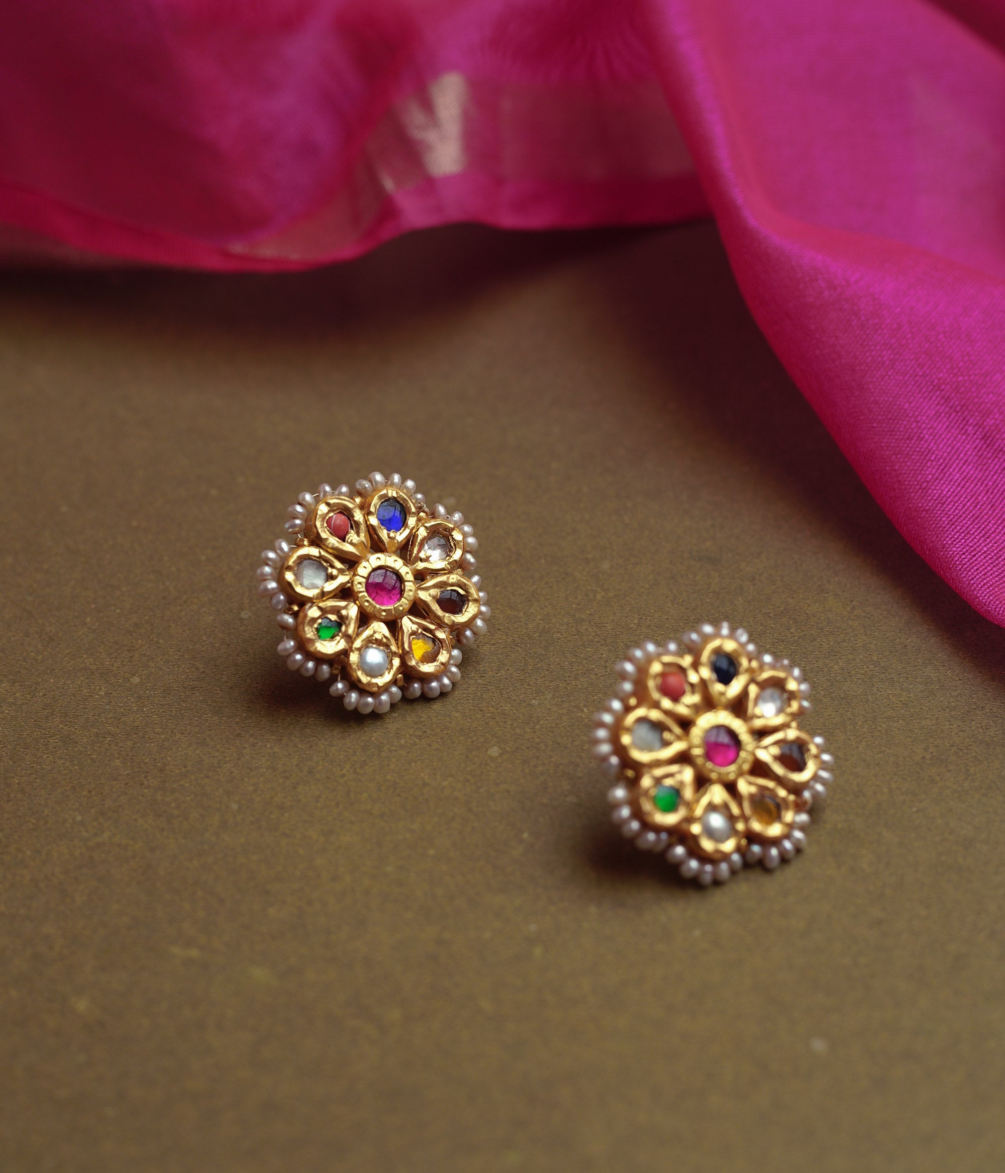 Close-up of two ornate gold stud earrings. Each features a central red gem surrounded by colorful Navratna stones and an outer border of small pearls, resting on a brown surface.
