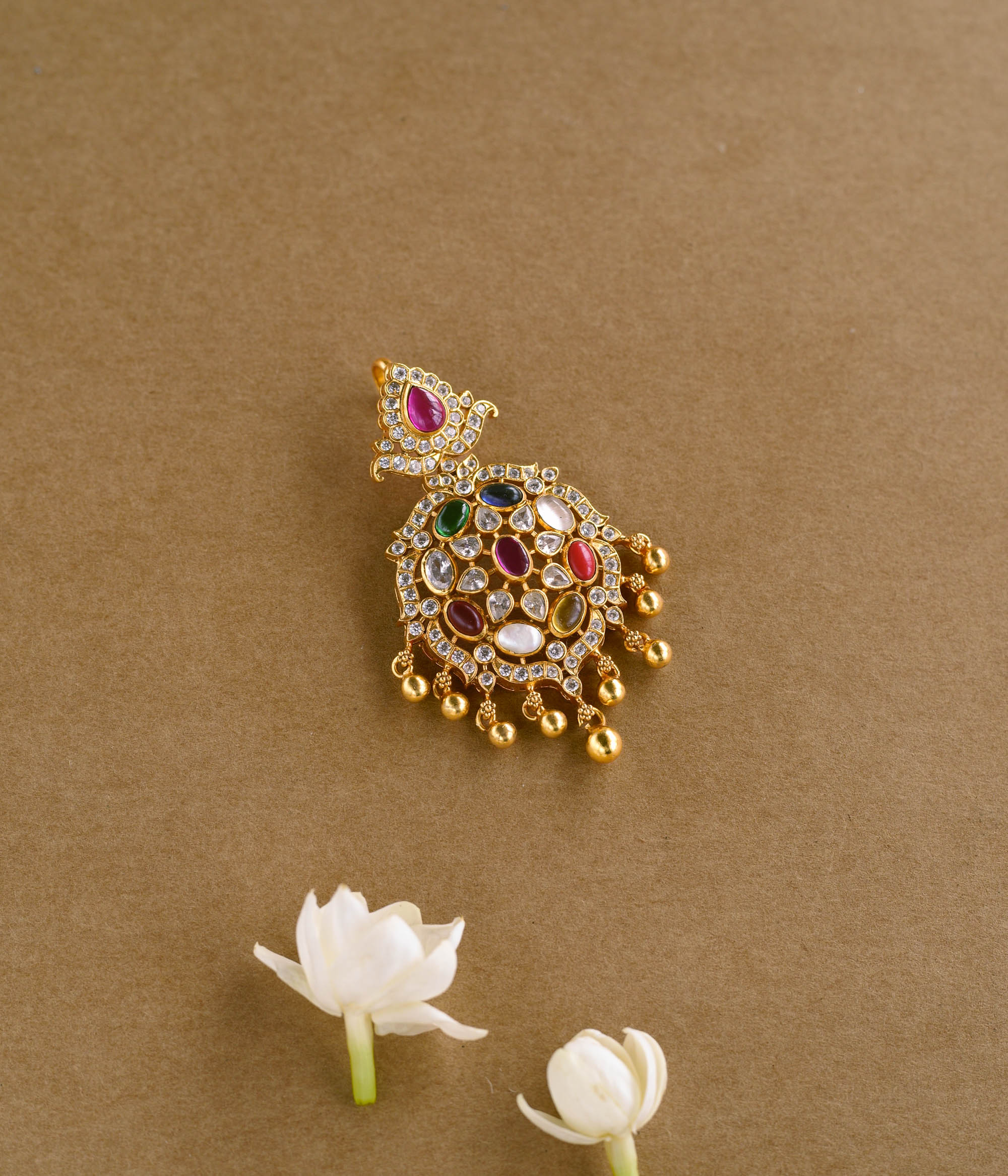 A gold pendant with an ornate, asymmetrical design. It is adorned with various colorful Navratna stones, clear stones, white Polki-like stones, and a single red stone at the top. Small gold beads dangle from the bottom.