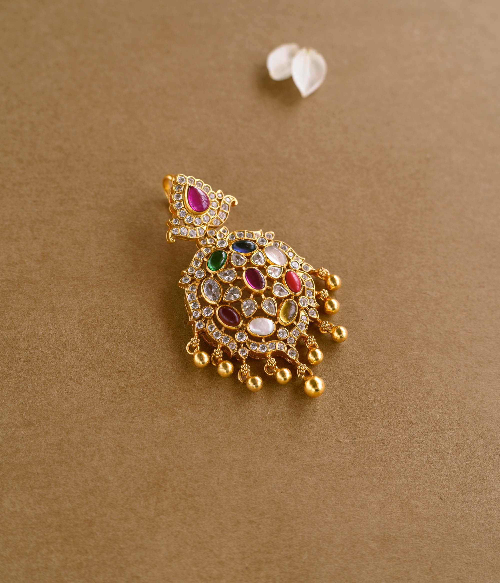 A gold pendant with an ornate, asymmetrical design. It is adorned with various colorful Navratna stones, clear stones, white Polki-like stones, and a single red stone at the top. Small gold beads dangle from the bottom.