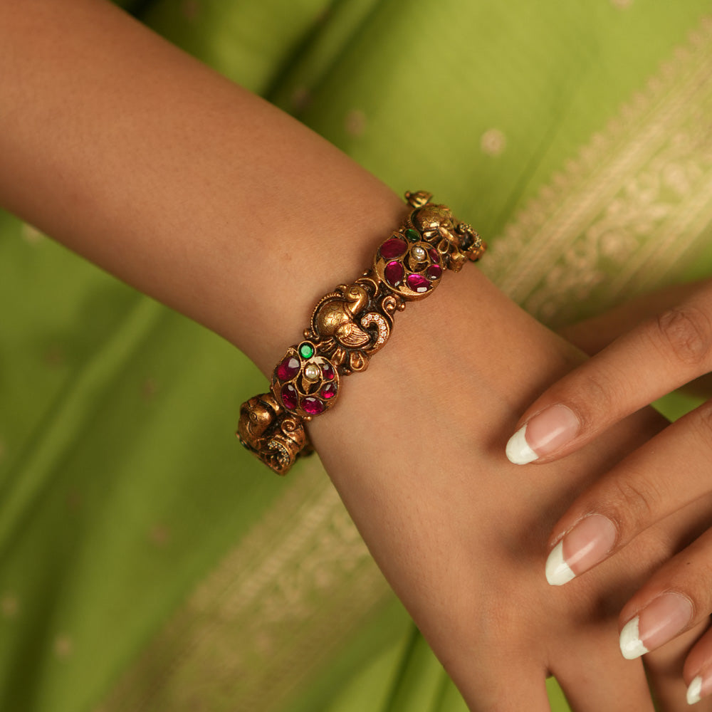 Regal whispers reborn in an antique gold-plated bangle, where peacock motifs and vibrant kundan stones echo timeless heritage and artistry.
