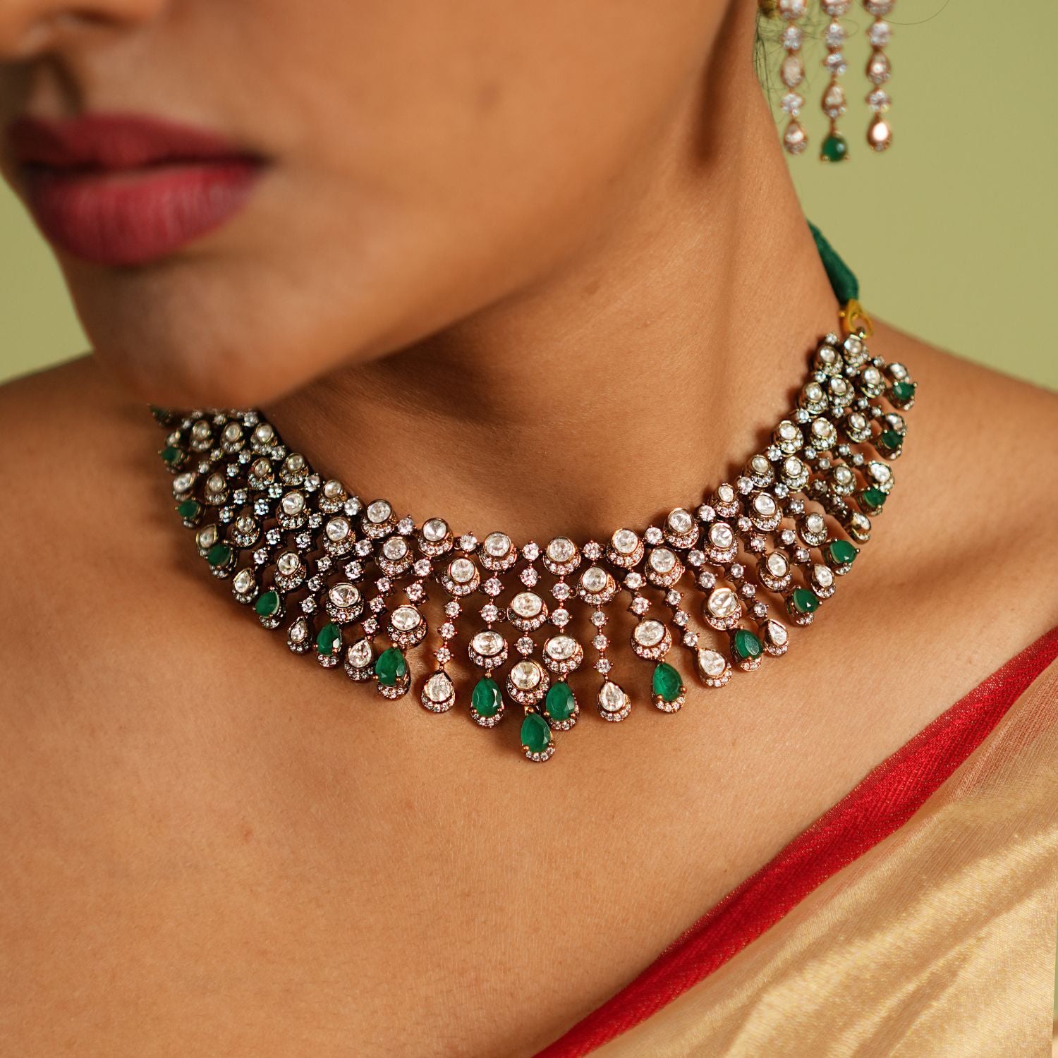This Victorian-inspired short necklace showcases cascading Polki stones, green gemstones, and CZ stones, creating a mesmerizing display of elegance. Each delicate drop catches the light, casting a spell of sophistication and timeless charm on the wearer.