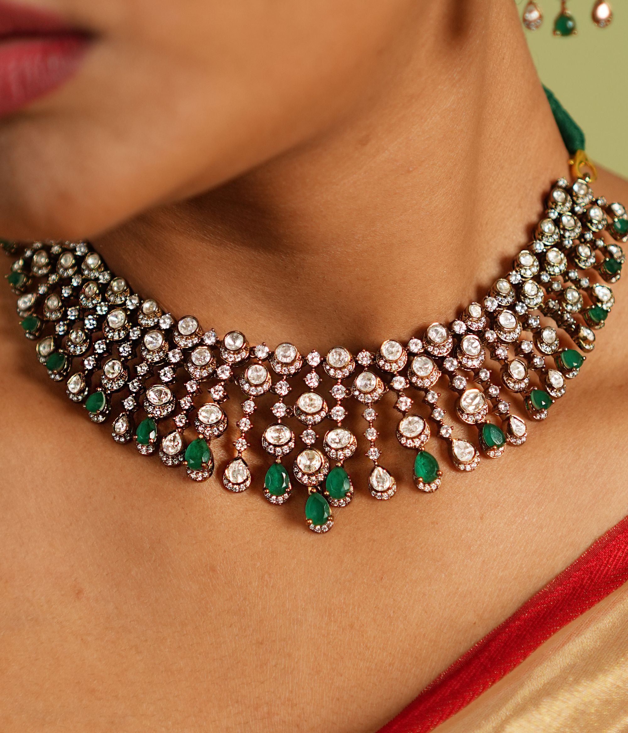 This Victorian-inspired short necklace showcases cascading Polki stones, green gemstones, and CZ stones, creating a mesmerizing display of elegance. Each delicate drop catches the light, casting a spell of sophistication and timeless charm on the wearer.