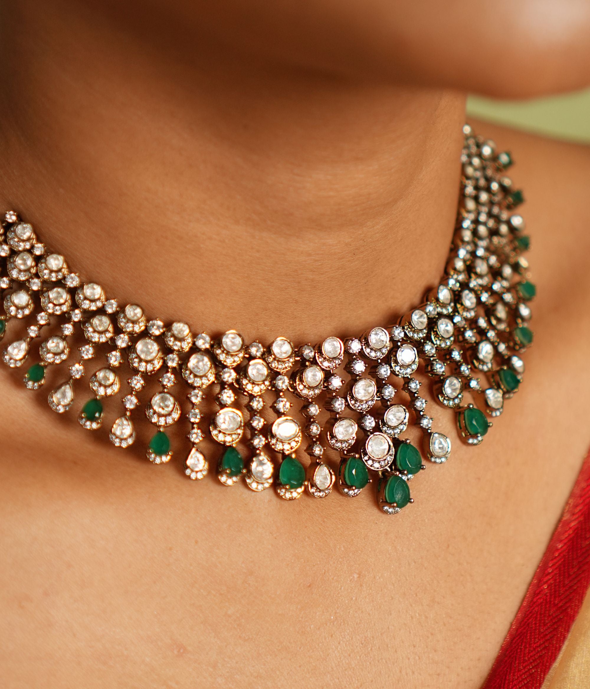 This Victorian-inspired short necklace showcases cascading Polki stones, green gemstones, and CZ stones, creating a mesmerizing display of elegance. Each delicate drop catches the light, casting a spell of sophistication and timeless charm on the wearer.