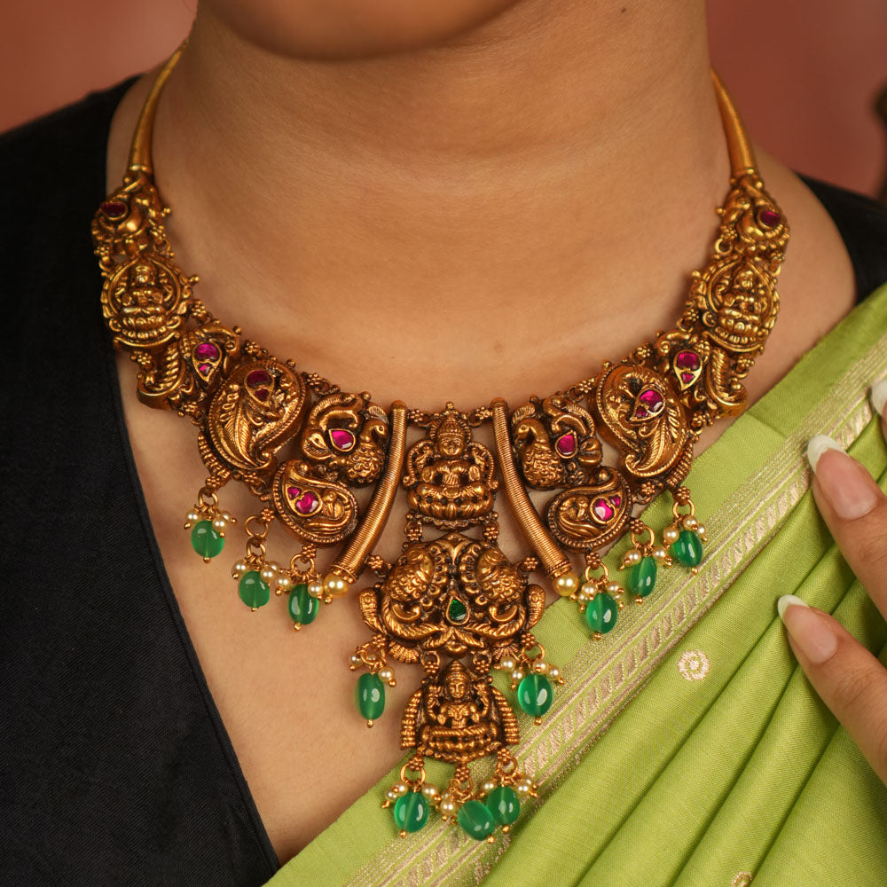 Rooted in ancient splendor, this antique gold-plated temple necklace gleams with goddess motifs, red kundan, pearls, and cascading green beads.