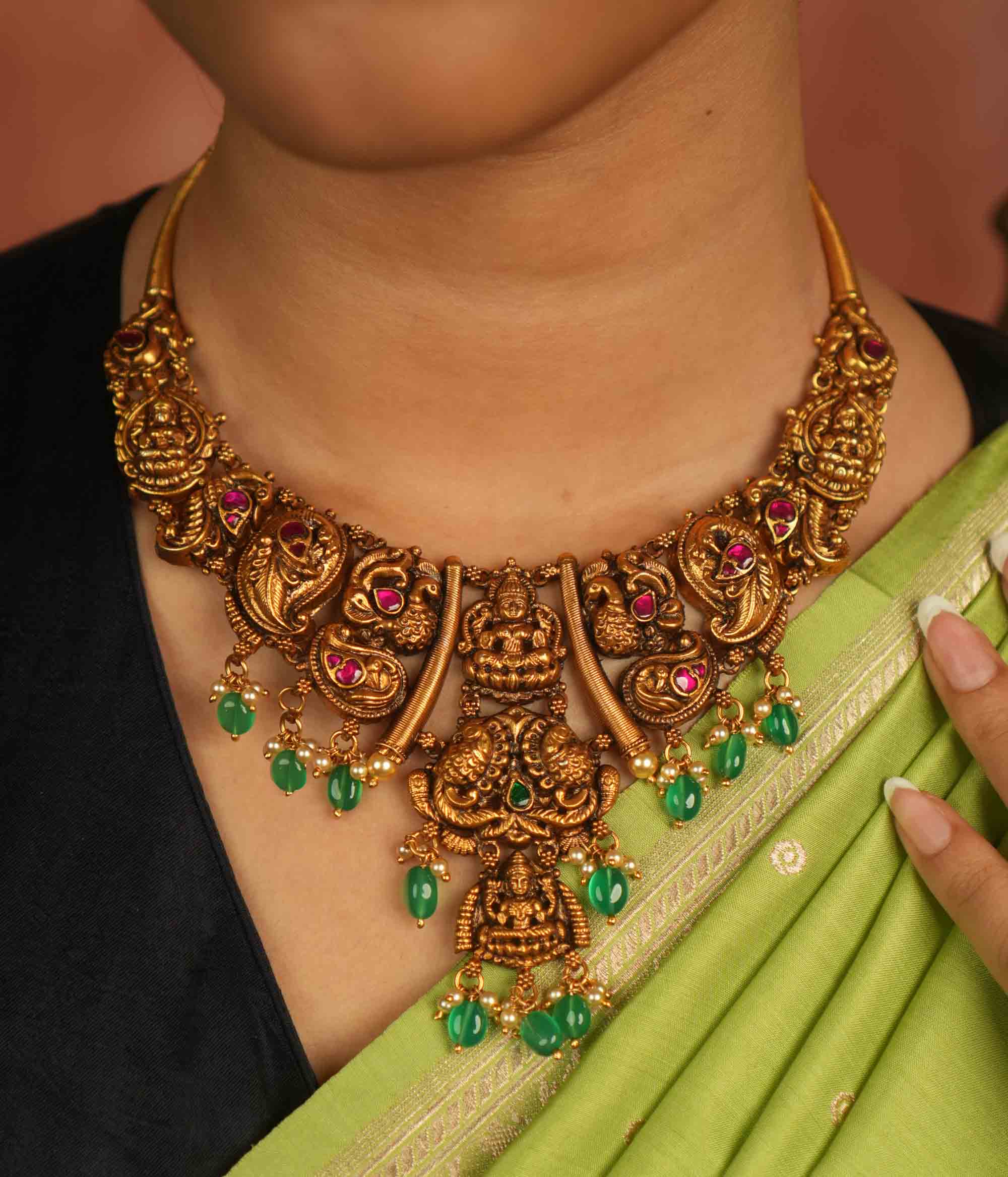 Rooted in ancient splendor, this antique gold-plated temple necklace gleams with goddess motifs, red kundan, pearls, and cascading green beads.
