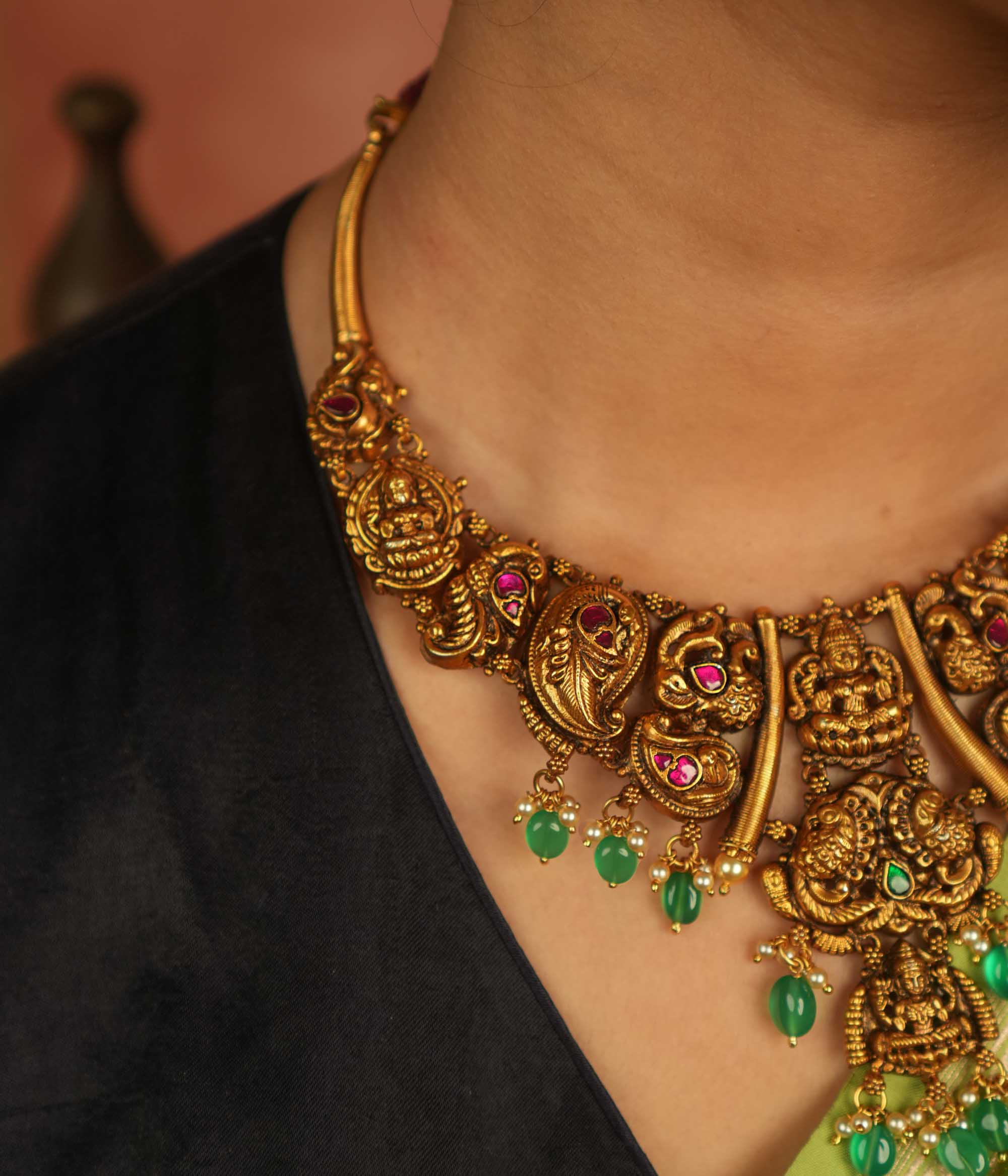 Rooted in ancient splendor, this antique gold-plated temple necklace gleams with goddess motifs, red kundan, pearls, and cascading green beads.
