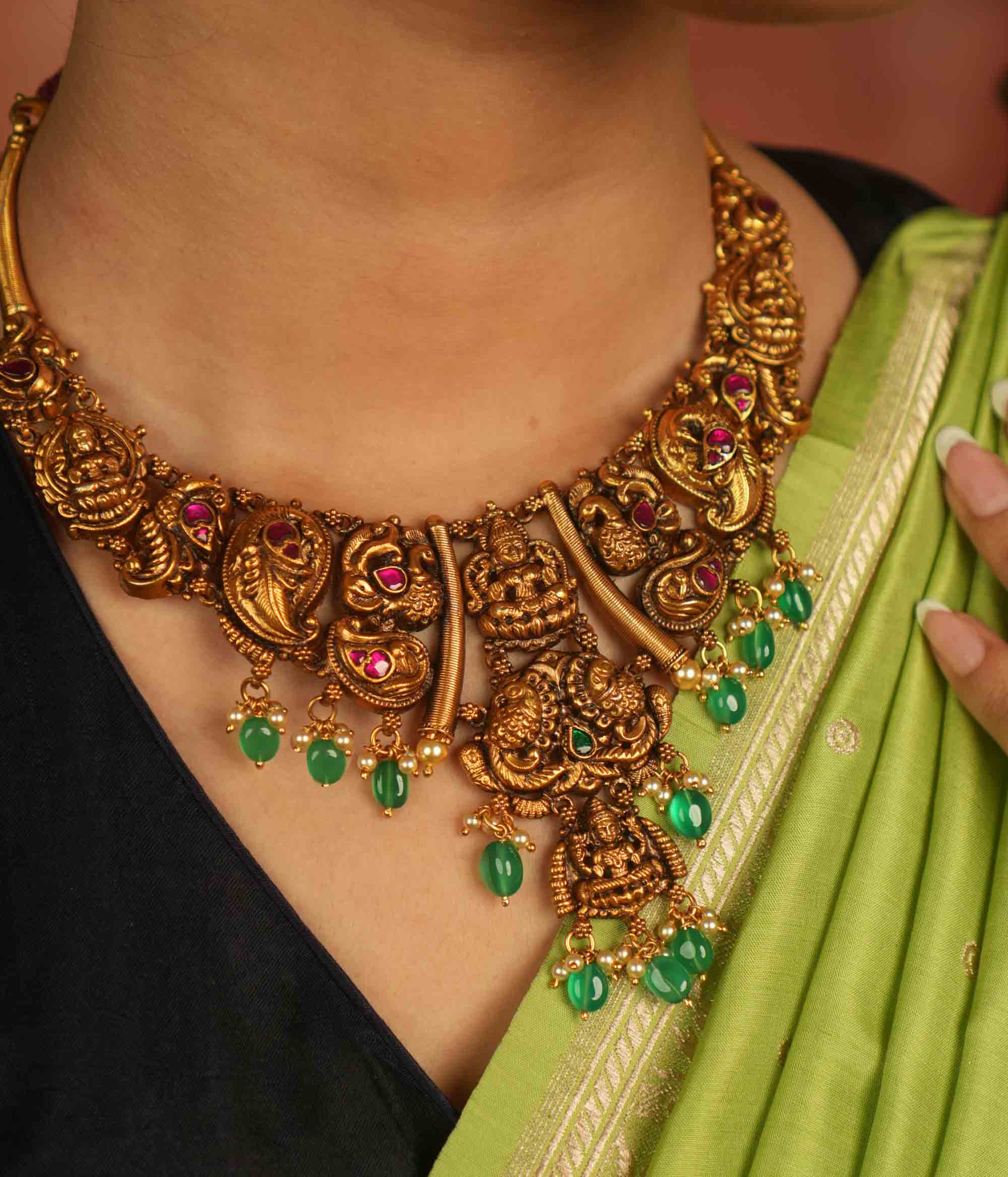 Rooted in ancient splendor, this antique gold-plated temple necklace gleams with goddess motifs, red kundan, pearls, and cascading green beads.