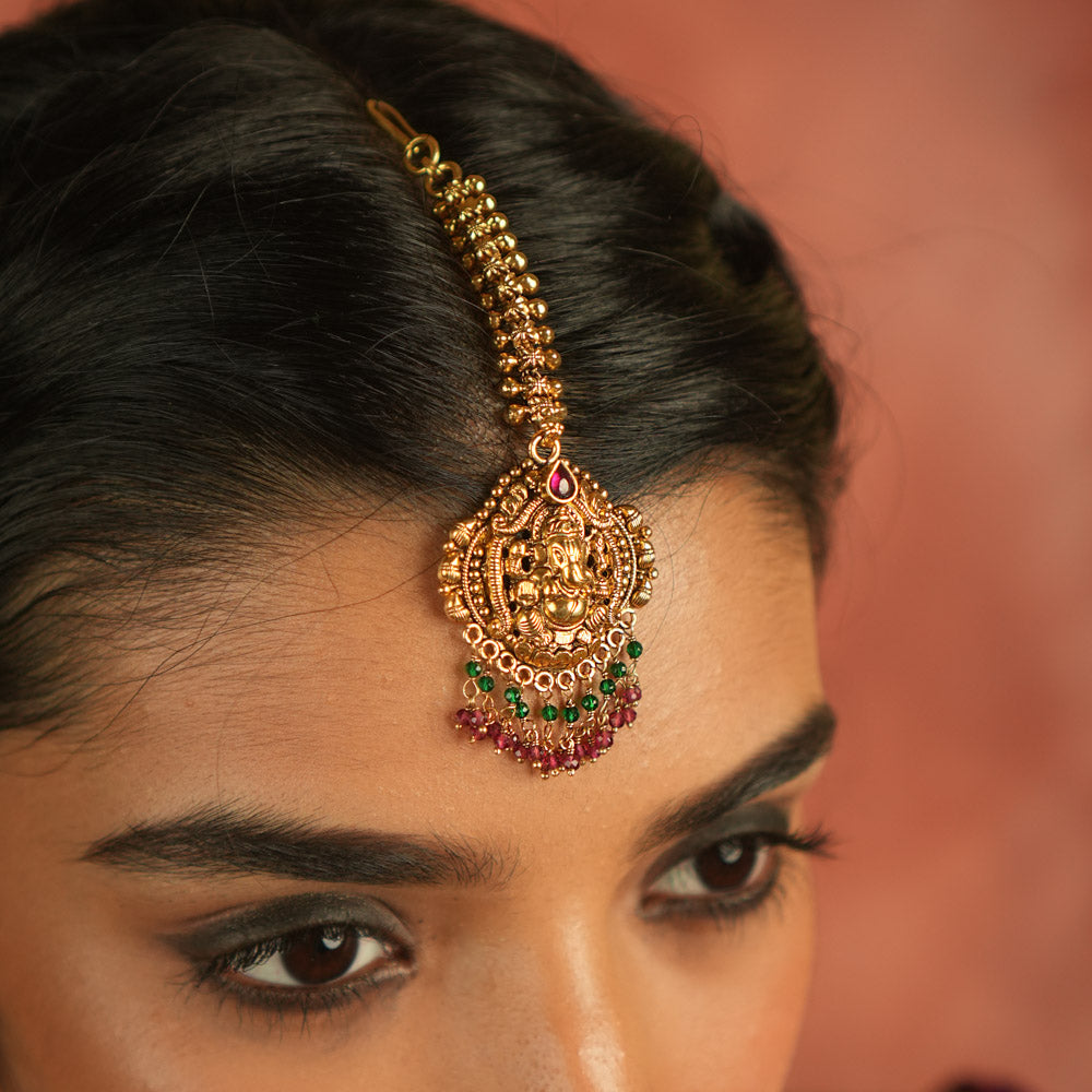 Sacred elegance reimagined! This gold-plated maangtika, adorned with a Lord Ganesha motif, glows with red spinel stones and beads, embodying tradition and spiritual grace.