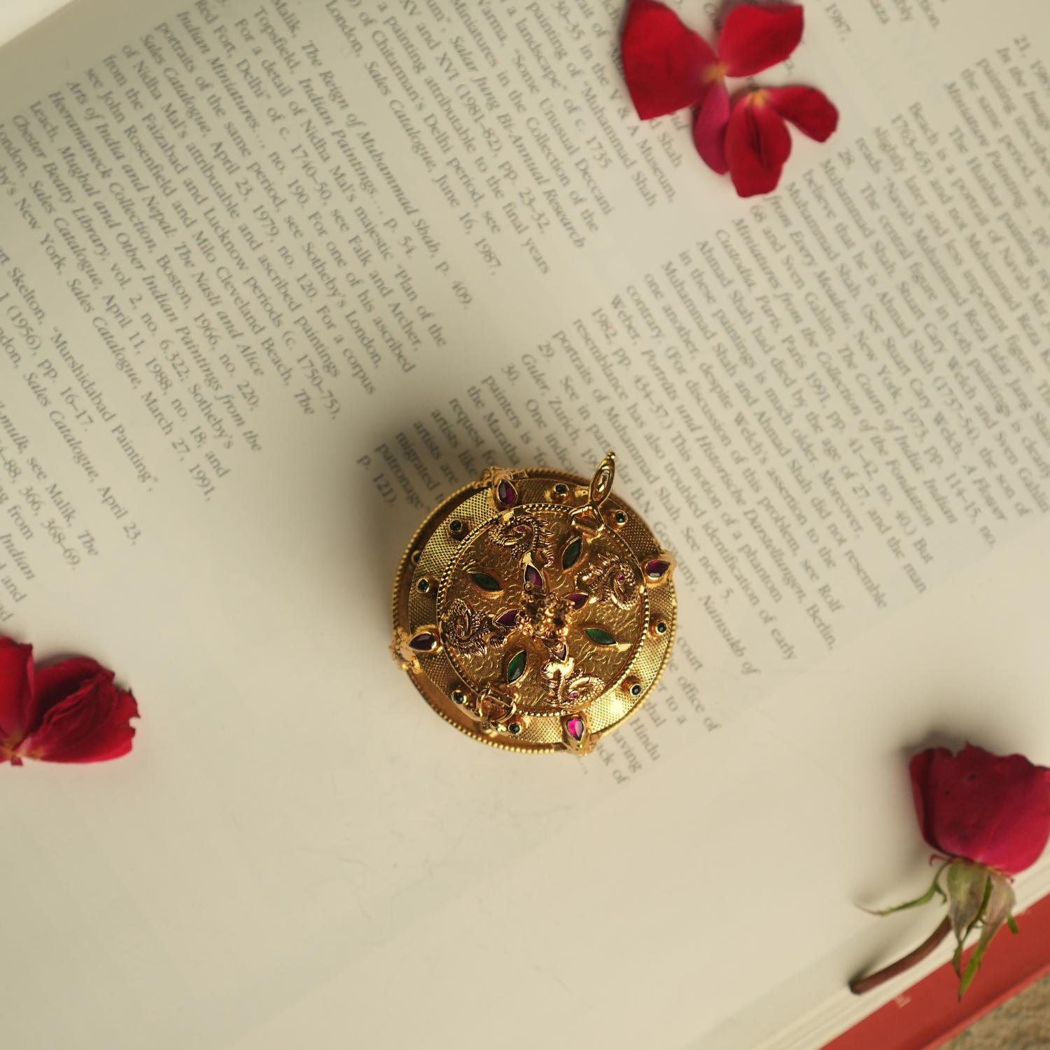 An intricately crafted antique gold-plated silver sindoor case, adorned with temple motifs and red and green kundan stones, embodies the essence of sacred elegance and timeless beauty.