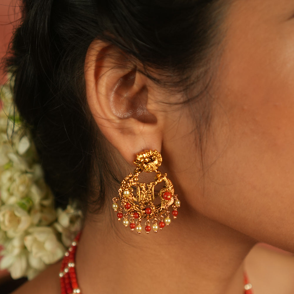 Timeless devotion reimagined! These gold-plated Radha Krishna dangler earrings, framed in an elegant peacock design, shimmer with red beads, embodying divine love and grace.