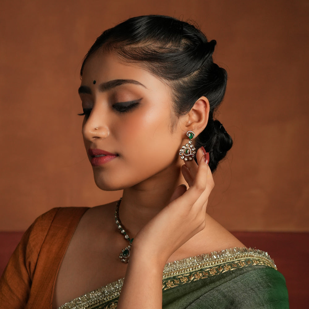 A symphony of Victorian and Oriental charm, these gold-plated silver earrings feature delicate polki and green gemstones. Their floral design, with a lush emerald center encircled by shimmering polki and CZ stones, whispers tales of timeless beauty. Intricate craftsmanship meets regal allure, creating an enchanting statement of elegance.