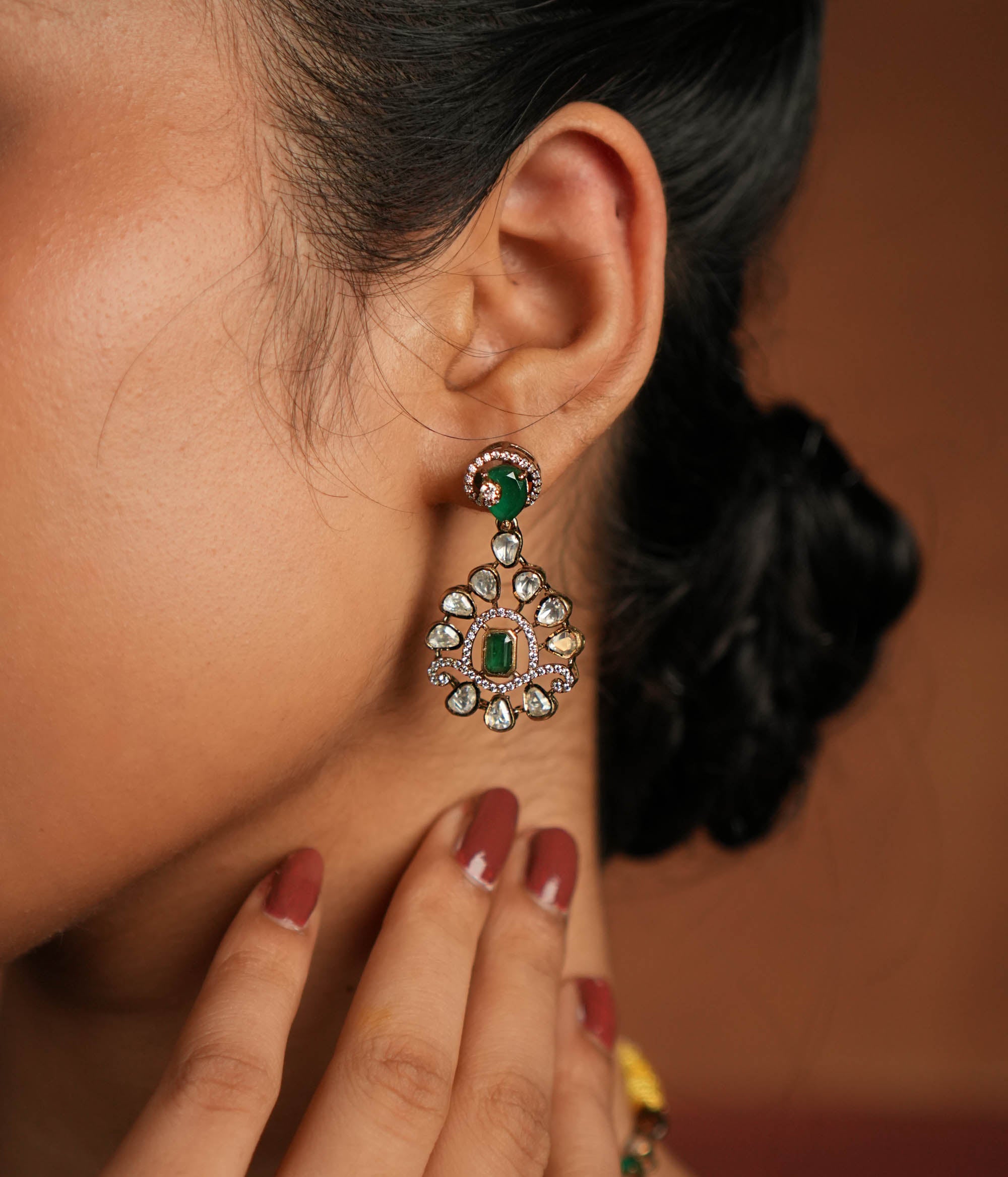 A symphony of Victorian and Oriental charm, these gold-plated silver earrings feature delicate polki and green gemstones. Their floral design, with a lush emerald center encircled by shimmering polki and CZ stones, whispers tales of timeless beauty. Intricate craftsmanship meets regal allure, creating an enchanting statement of elegance.