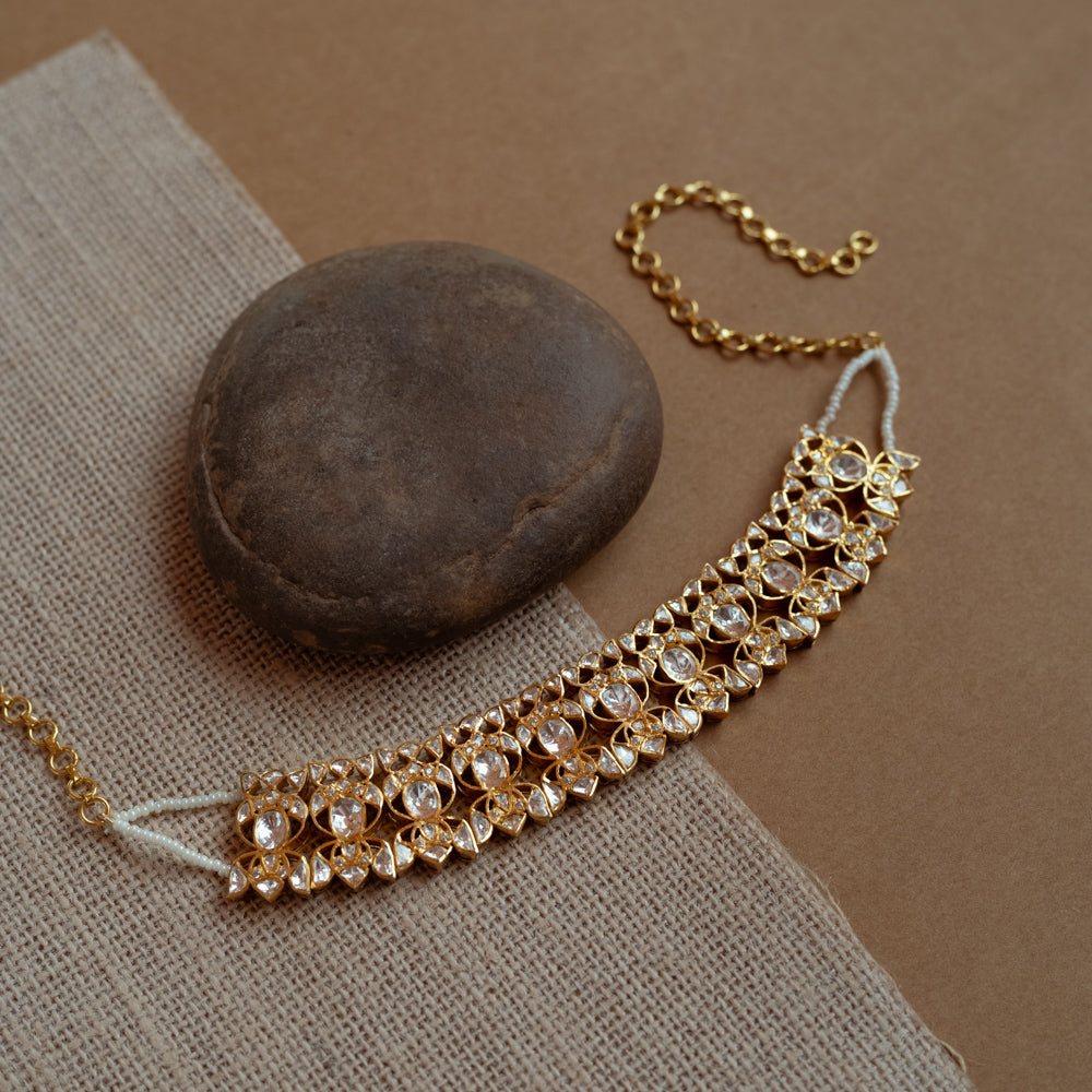 A delicate gold choker adorned with white Polki stones. It features an adjustable chain and rests on a dark stone and textured fabric.
