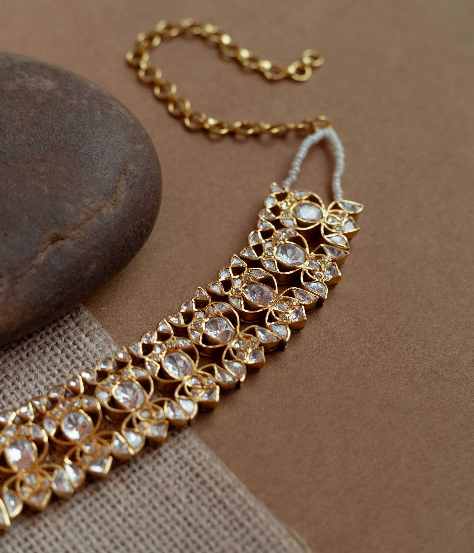 A delicate gold choker adorned with white Polki stones. It features an adjustable chain and rests on a dark stone and textured fabric.