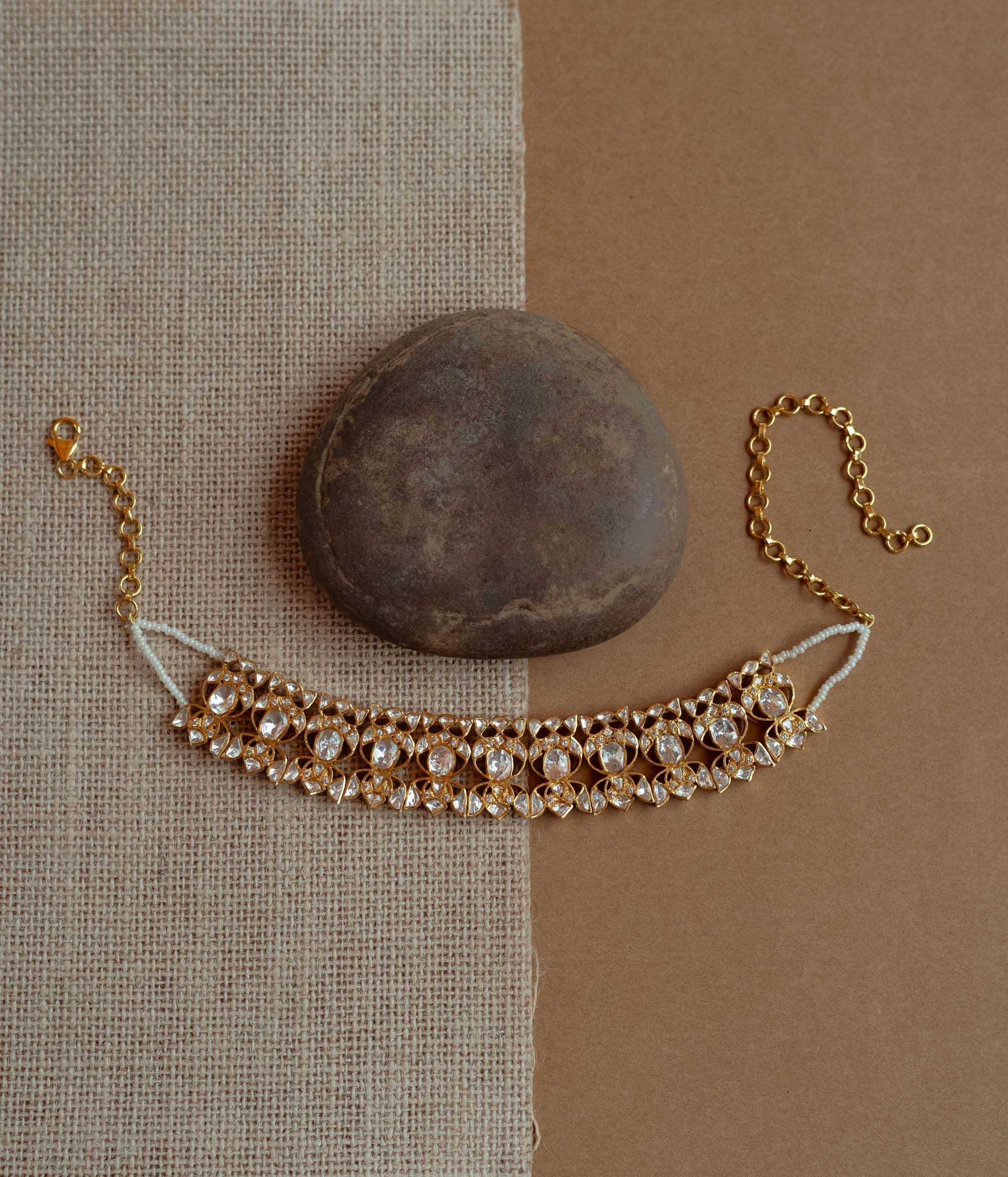 A delicate gold choker adorned with white Polki stones. It features an adjustable chain and rests on a dark stone and textured fabric.