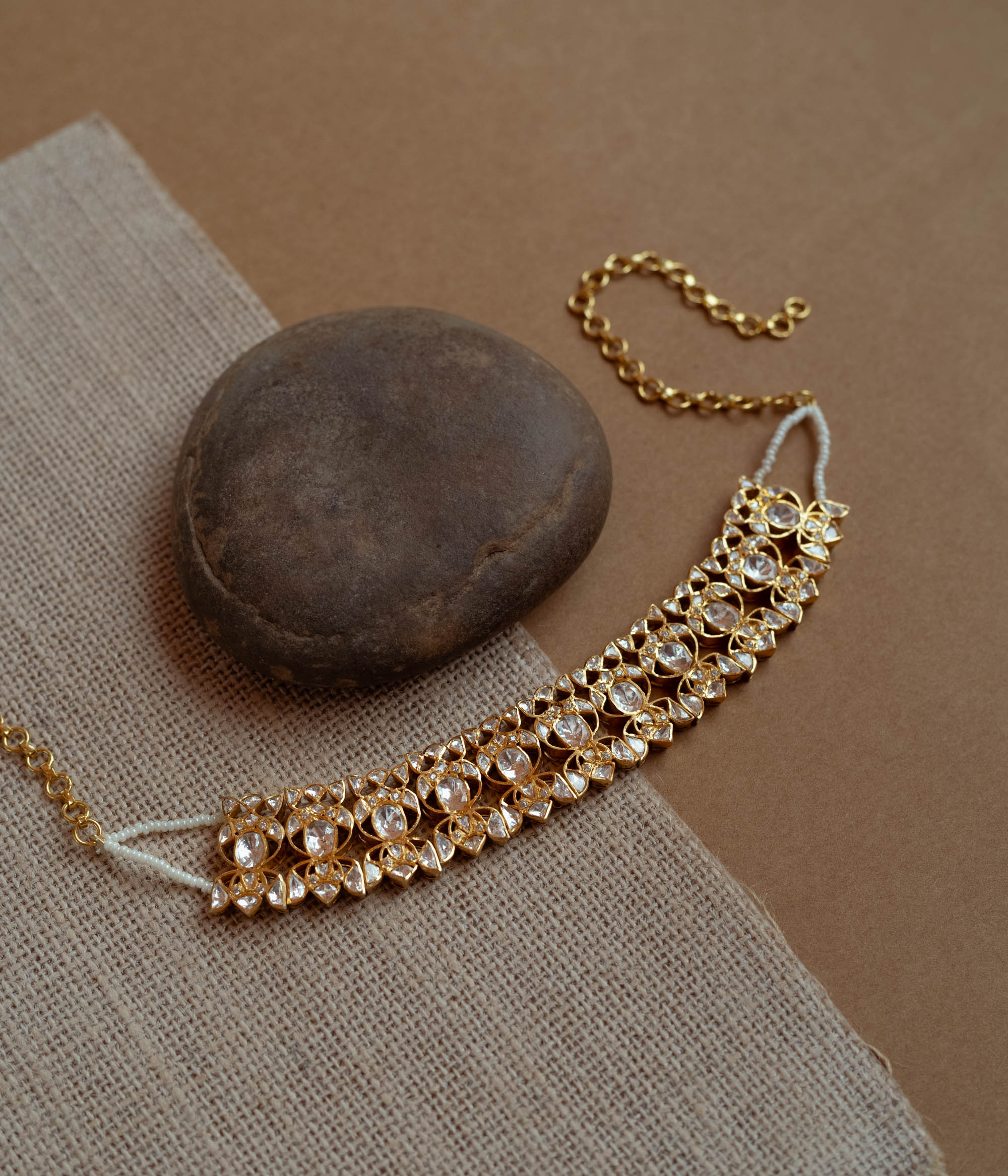 A delicate gold choker adorned with white Polki stones. It features an adjustable chain and rests on a dark stone and textured fabric.