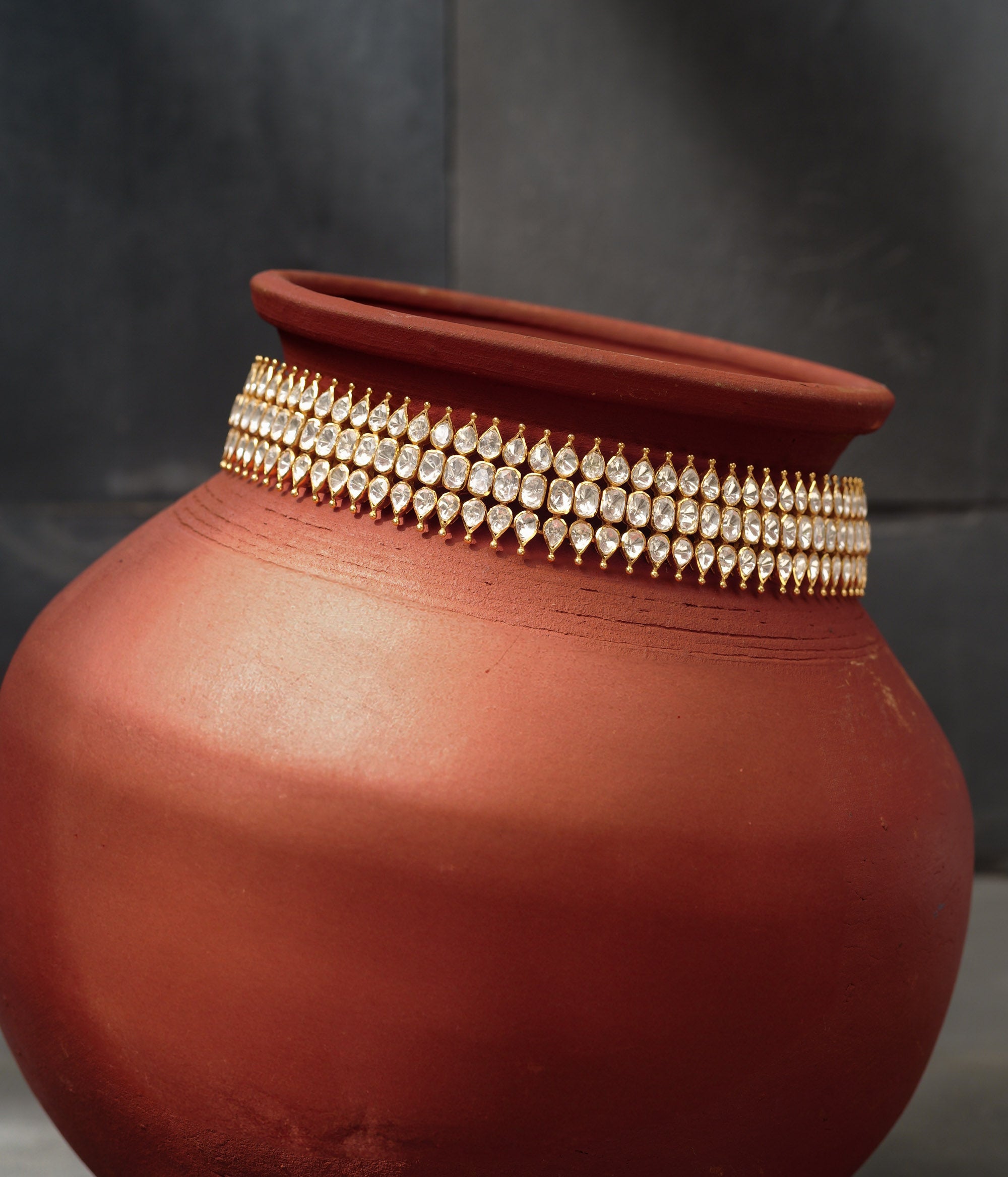 Close-up of a gold choker with three rows of teardrop-shaped white Polki stones. The choker is draped around a terracotta pot.