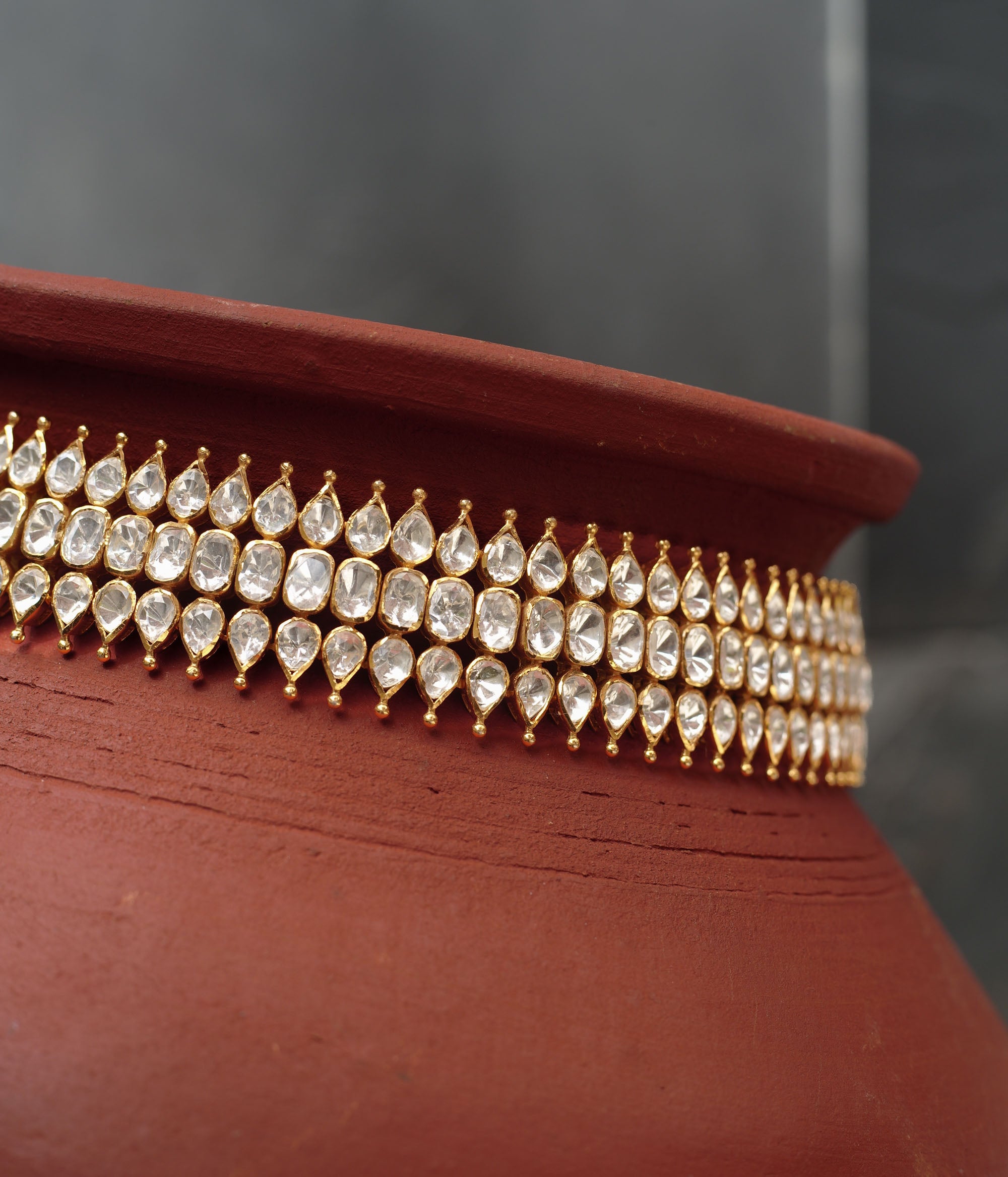 Close-up of a gold choker with three rows of teardrop-shaped white Polki stones. The choker is draped around a terracotta pot.