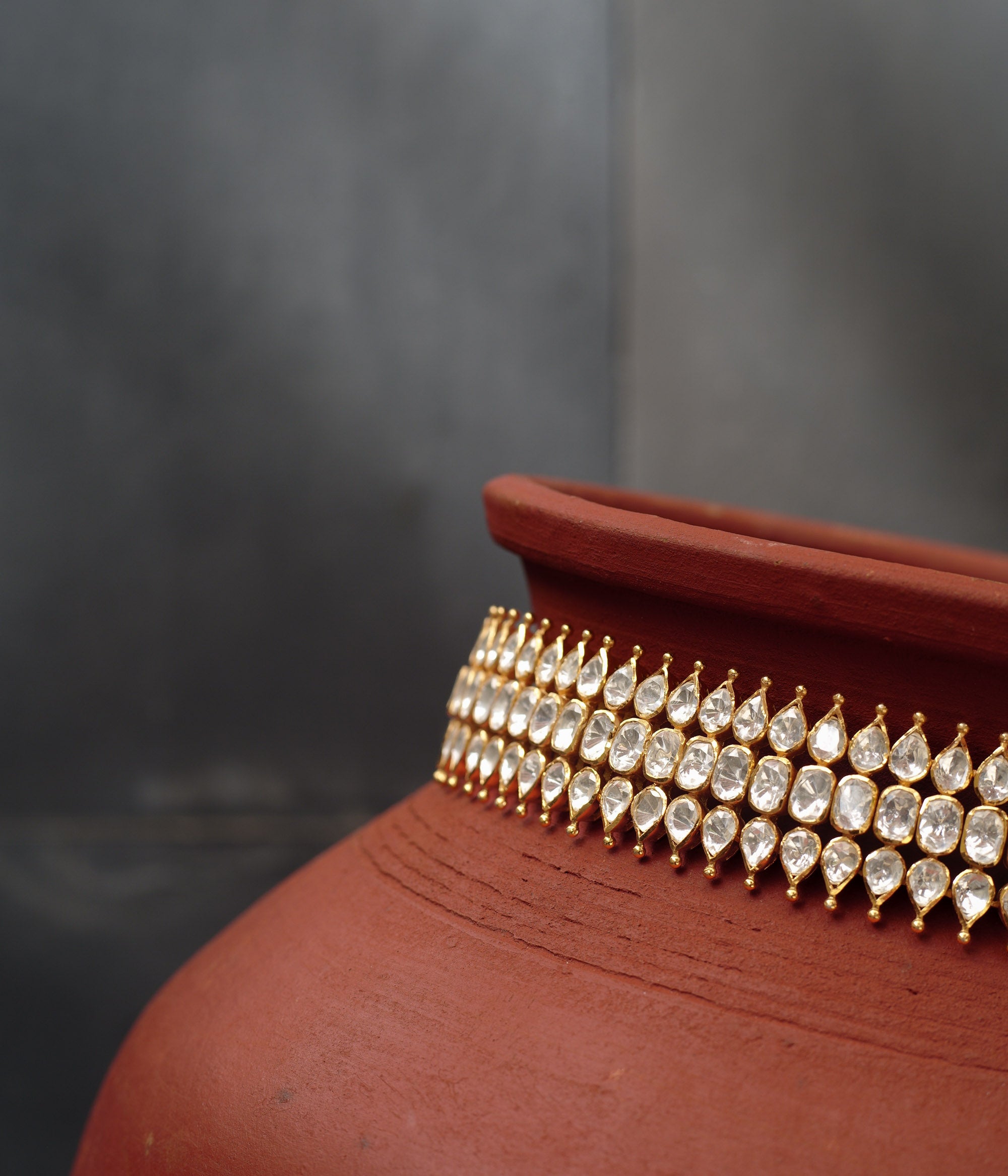 Close-up of a gold choker with three rows of teardrop-shaped white Polki stones. The choker is draped around a terracotta pot.