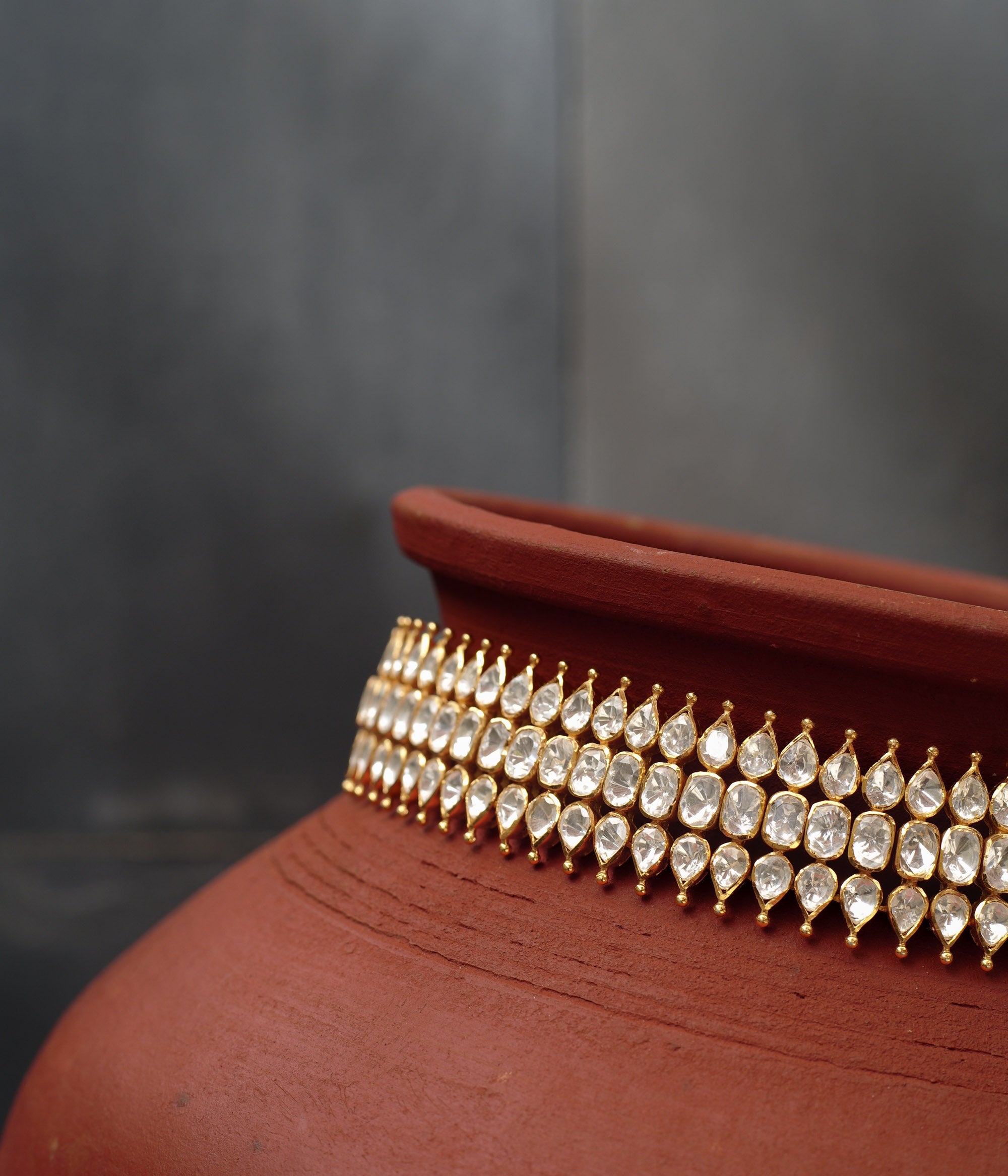 Close-up of a gold choker with three rows of teardrop-shaped white Polki stones. The choker is draped around a terracotta pot.
