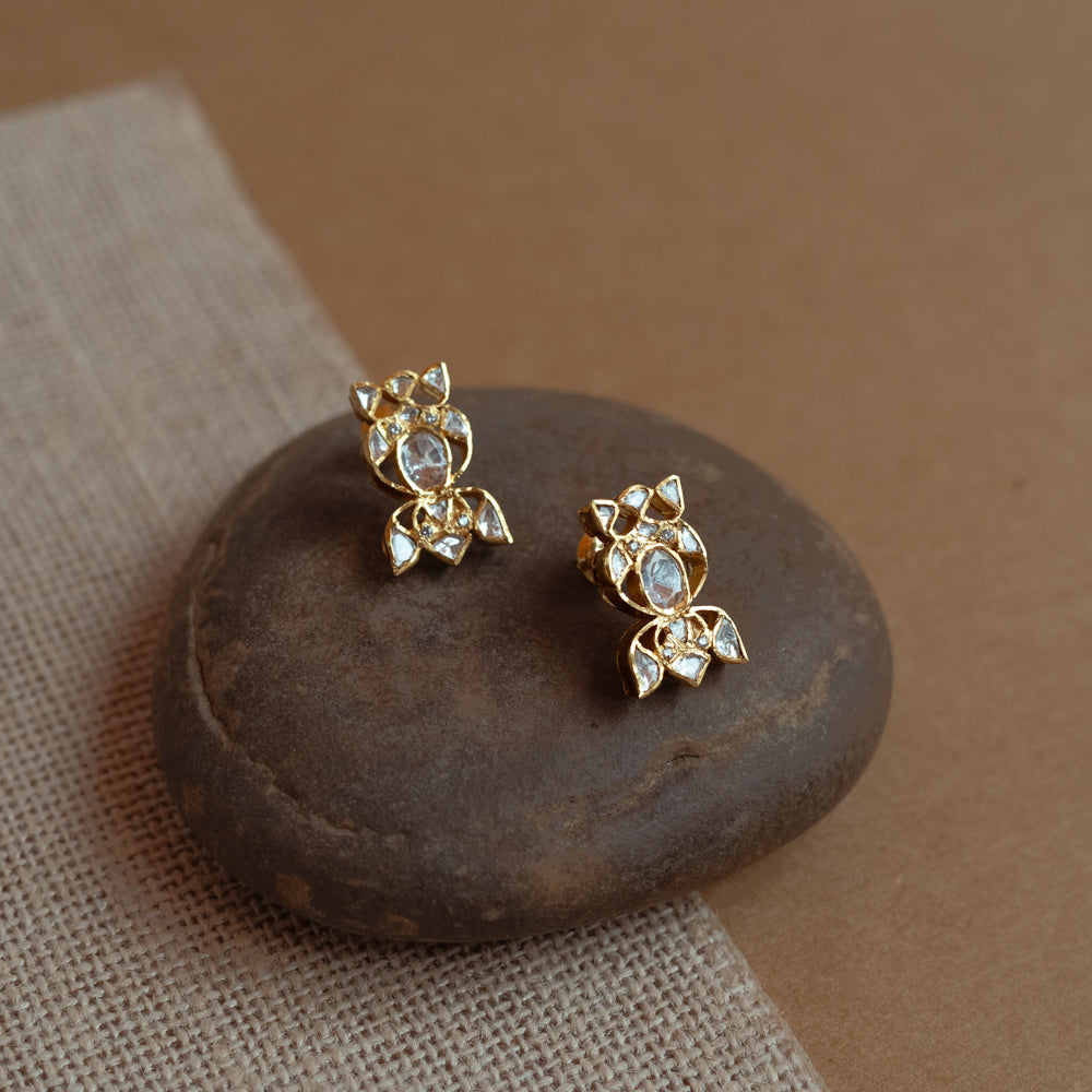 Close-up of two delicate gold stud earrings with a unique floral shape. Each is set with multiple small white Polki stones, resting on a smooth dark stone.