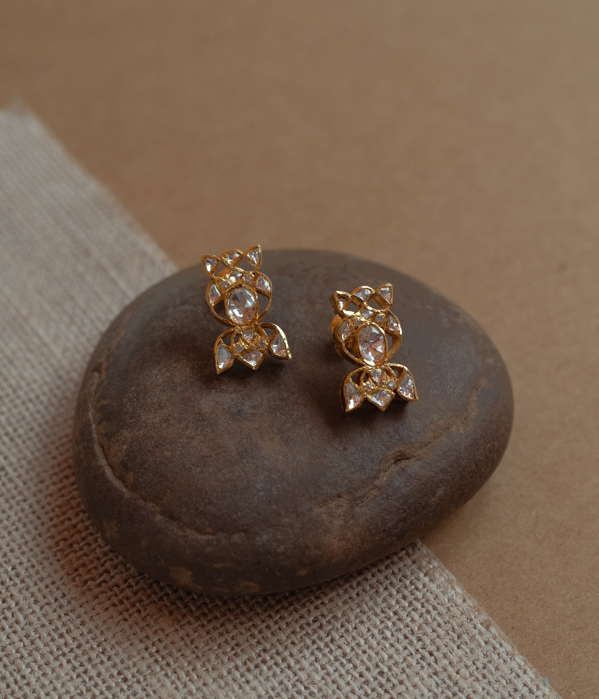 Close-up of two delicate gold stud earrings with a unique floral shape. Each is set with multiple small white Polki stones, resting on a smooth dark stone.