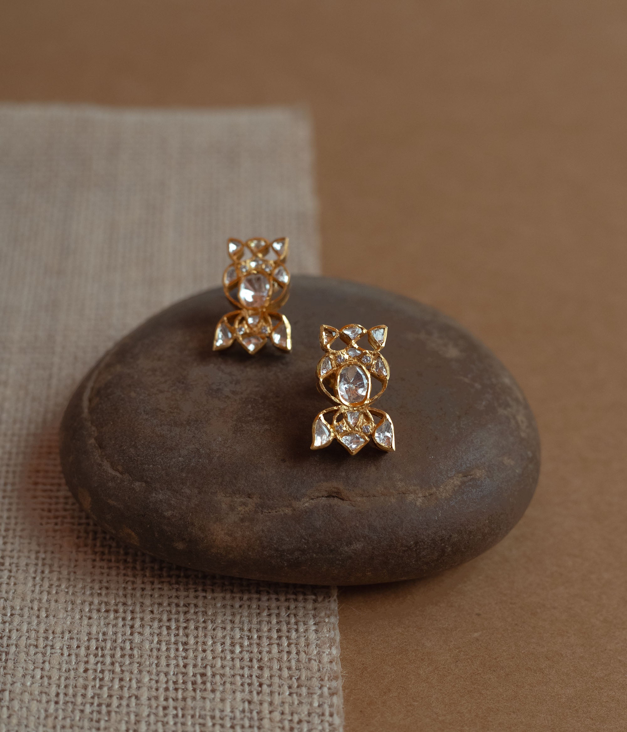 Close-up of two delicate gold stud earrings with a unique floral shape. Each is set with multiple small white Polki stones, resting on a smooth dark stone.