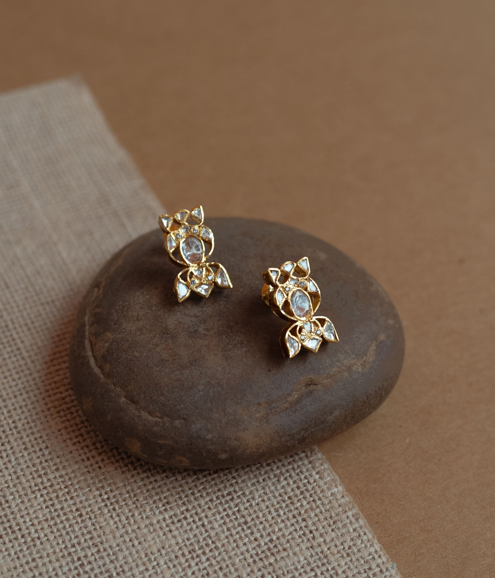 Close-up of two delicate gold stud earrings with a unique floral shape. Each is set with multiple small white Polki stones, resting on a smooth dark stone.