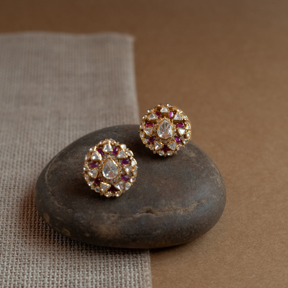 Close-up of two round gold stud earrings. Each features a central clear Polki stone surrounded by red Polki stones and smaller clear stones, resting on a smooth dark stone.