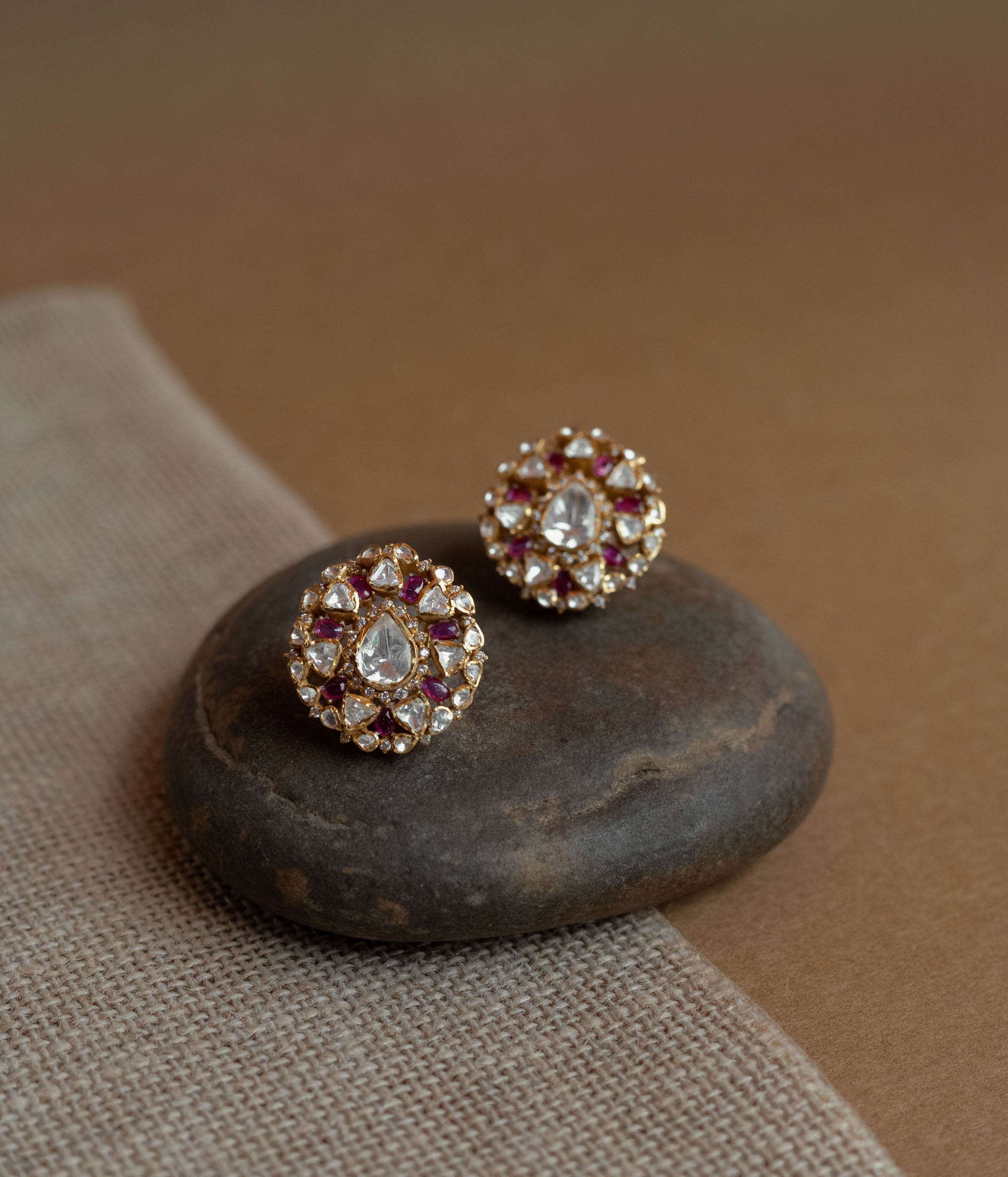 Close-up of two round gold stud earrings. Each features a central clear Polki stone surrounded by red Polki stones and smaller clear stones, resting on a smooth dark stone.