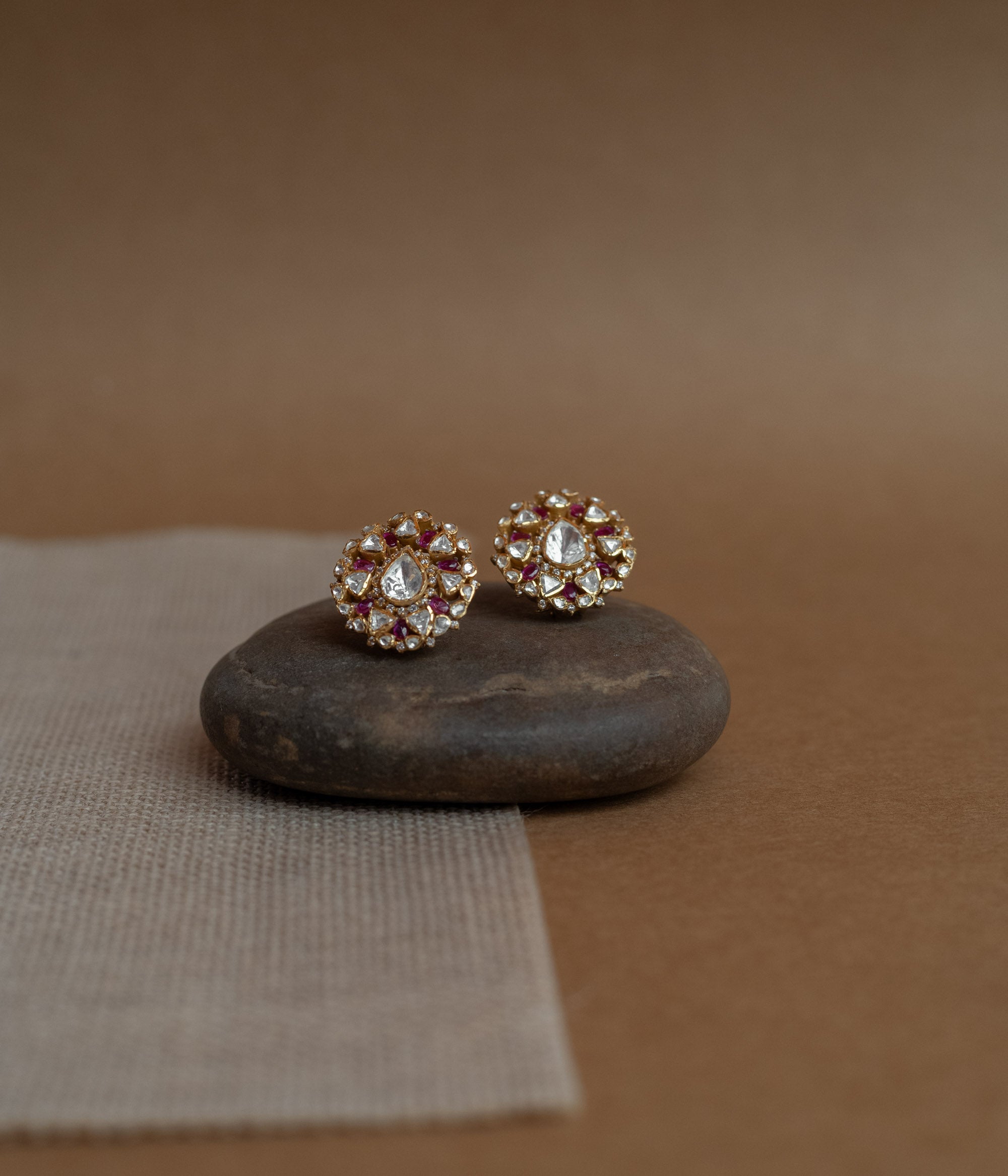 Close-up of two round gold stud earrings. Each features a central clear Polki stone surrounded by red Polki stones and smaller clear stones, resting on a smooth dark stone.