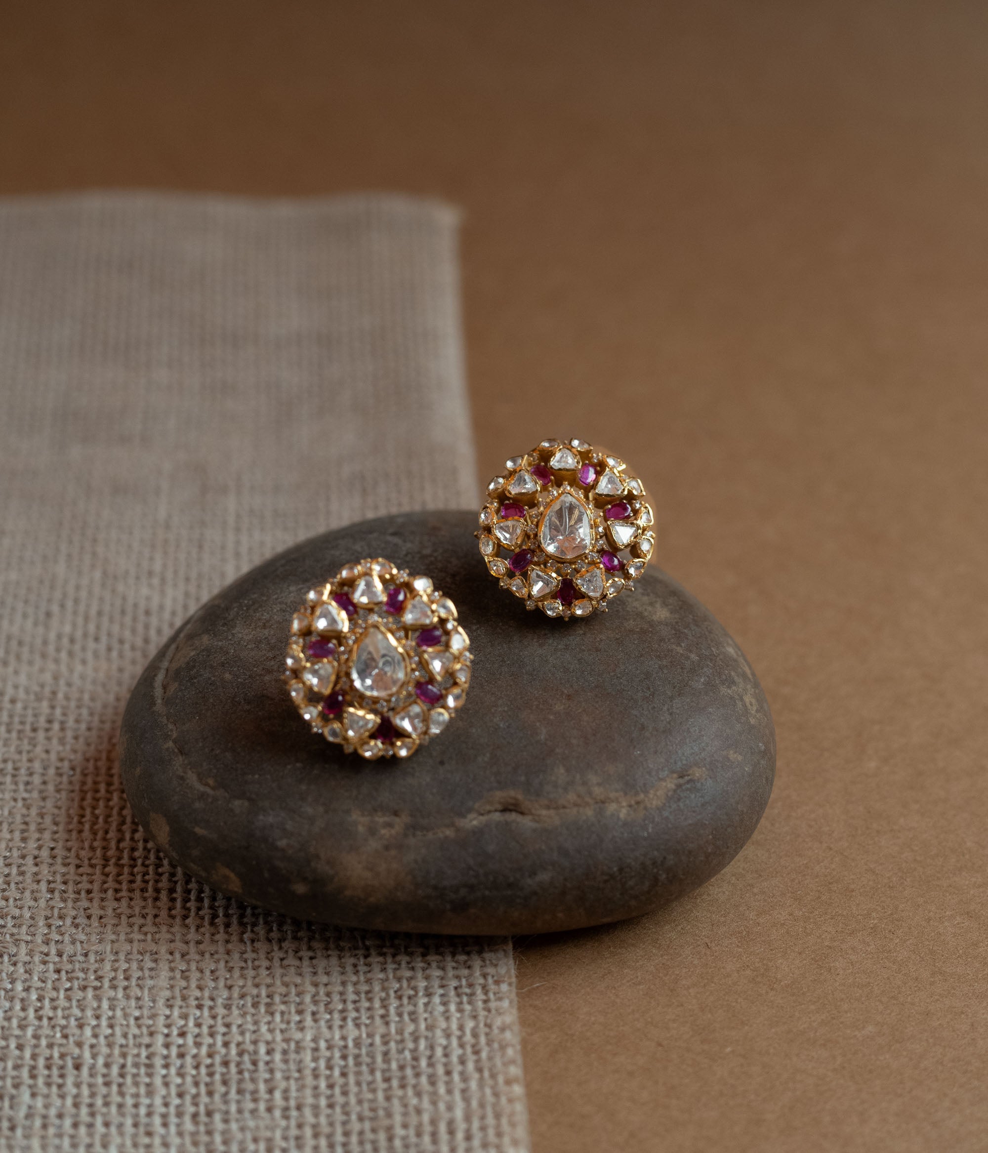 Close-up of two round gold stud earrings. Each features a central clear Polki stone surrounded by red Polki stones and smaller clear stones, resting on a smooth dark stone.