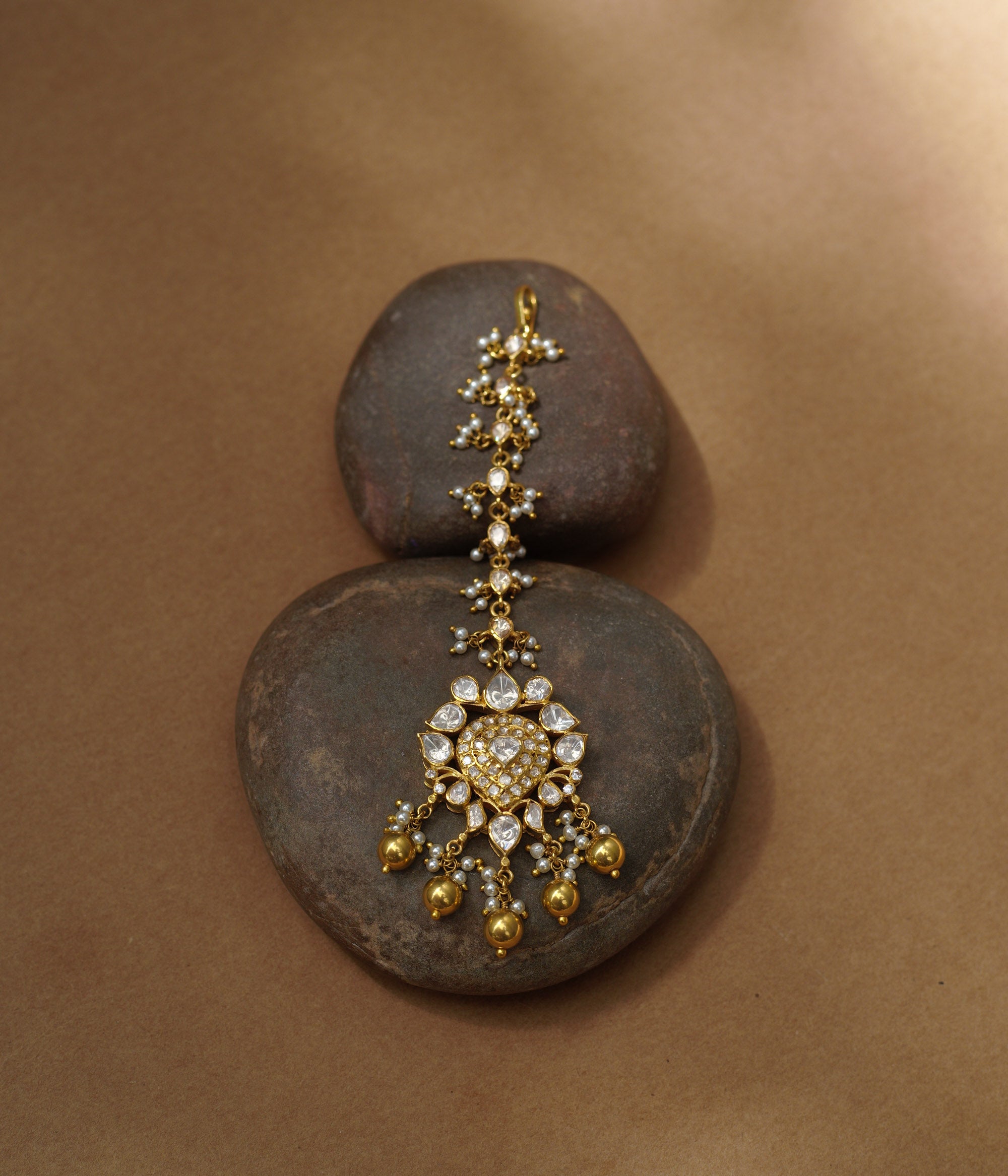 A golden maang tikka with a central heart-shaped pendant. It's adorned with white Polki stones and small dangling pearls, resting on two dark stones.