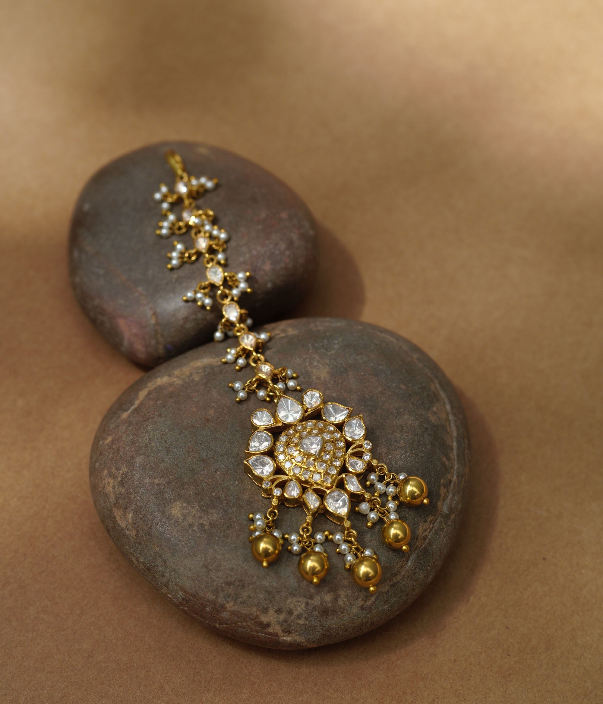 A golden maang tikka with a central heart-shaped pendant. It's adorned with white Polki stones and small dangling pearls, resting on two dark stones.