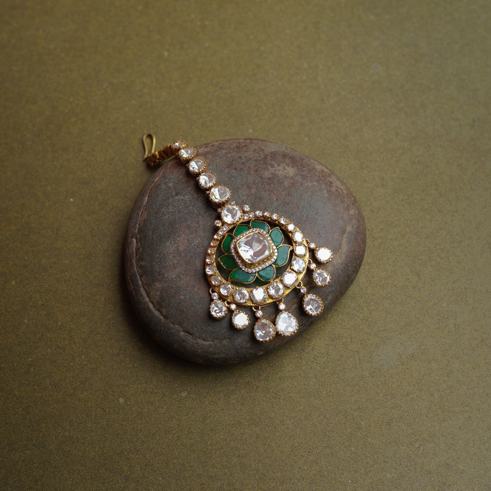 A golden maang tikka with a large round pendant. It features a prominent white Polki stone at its center, surrounded by green enamel, and dangling white Polki stones.