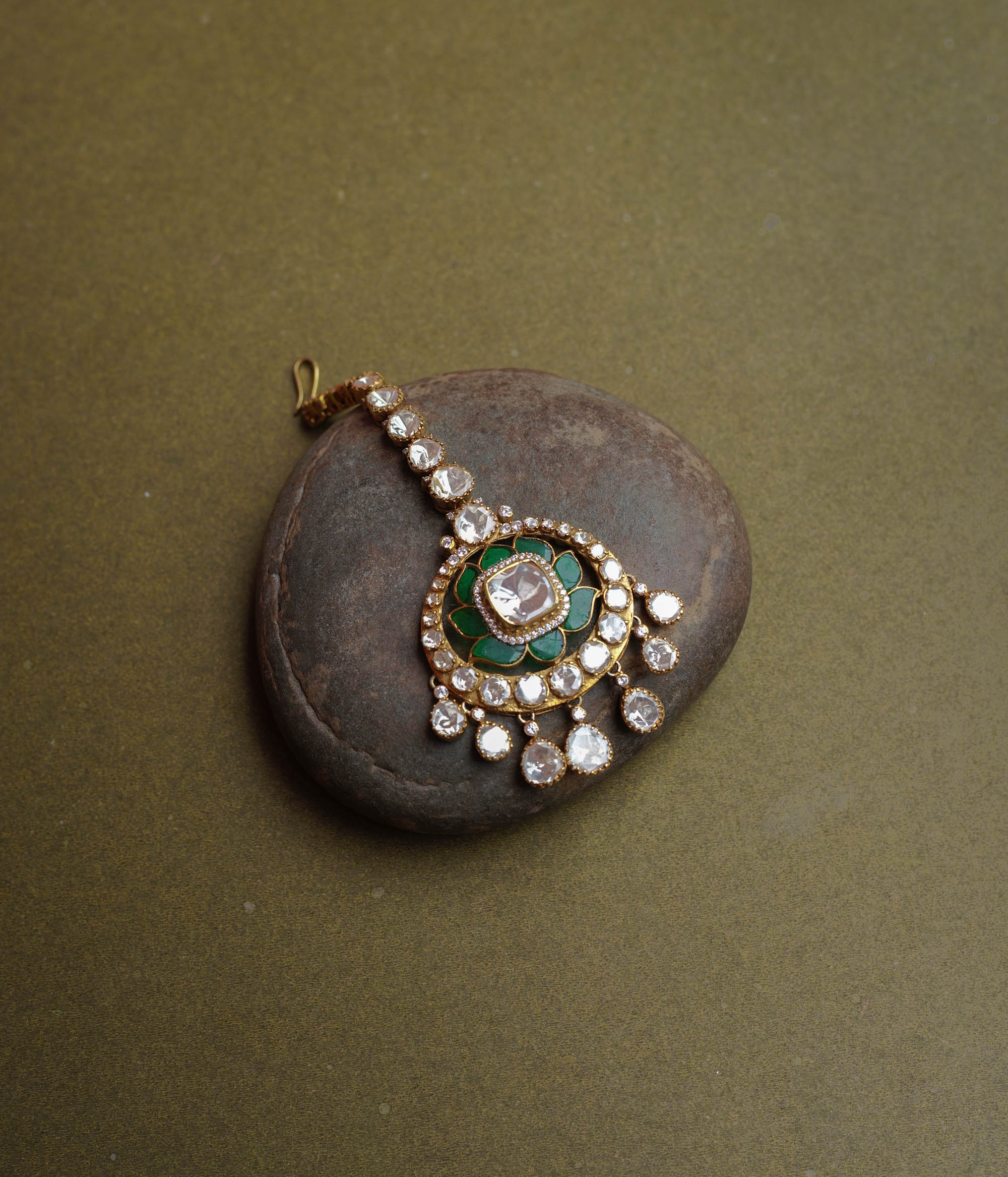 A golden maang tikka with a large round pendant. It features a prominent white Polki stone at its center, surrounded by green enamel, and dangling white Polki stones.