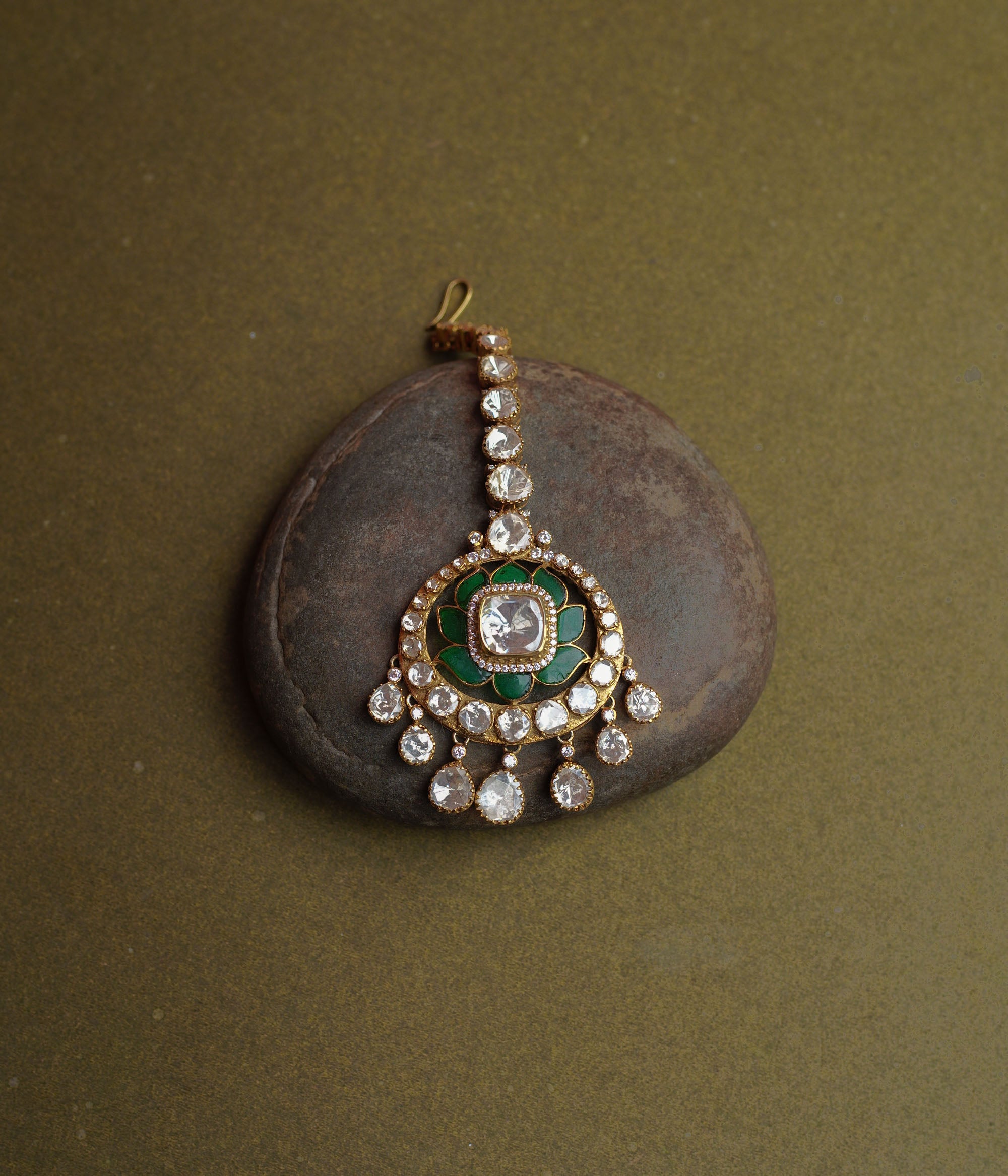 A golden maang tikka with a large round pendant. It features a prominent white Polki stone at its center, surrounded by green enamel, and dangling white Polki stones.