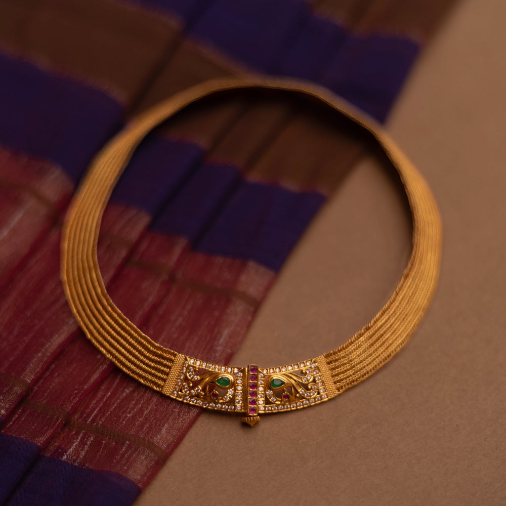 Close-up of a thick, golden choker necklace. It features intricate detailing with small green and clear stones at the clasp, set against a blurred fabric background.