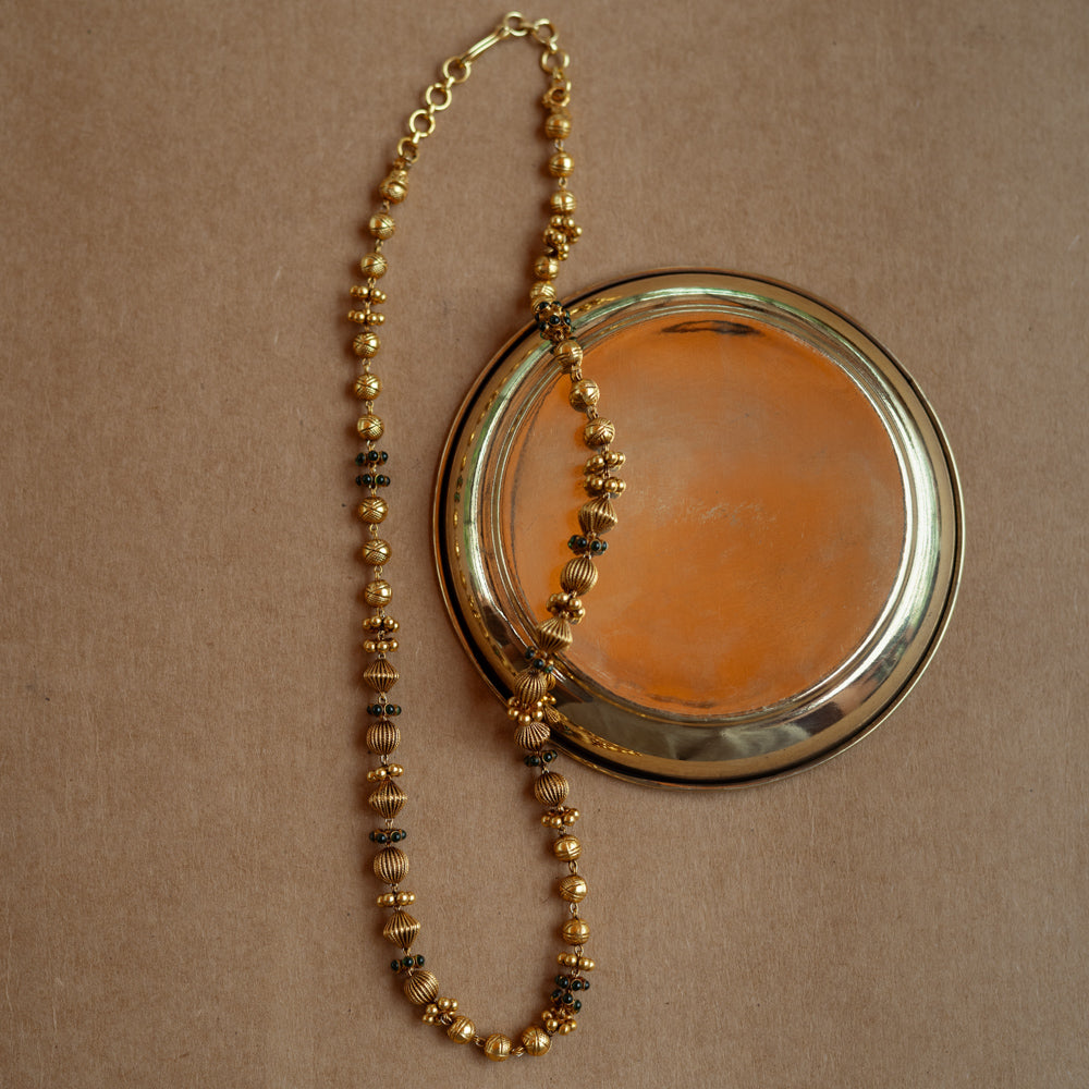 A long, delicate gold chain with various textured beads. Small green stones are interspersed along the chain, which is draped over a golden plate on a brown surface.