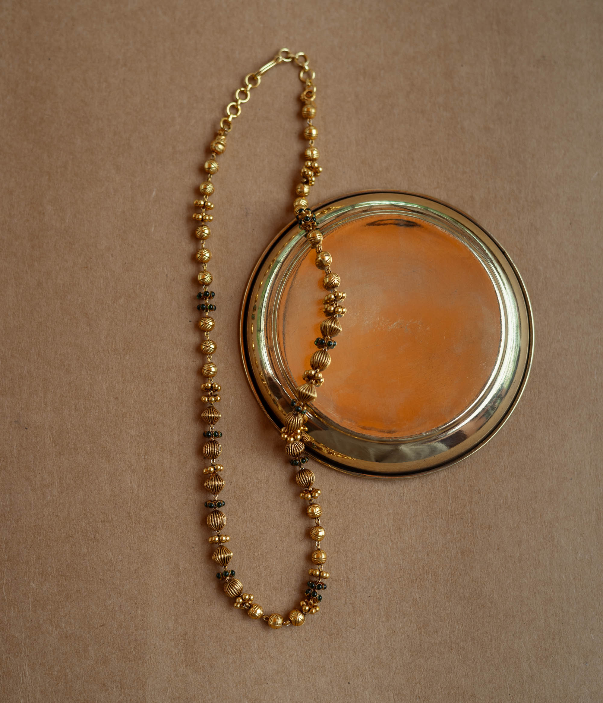 A long, delicate gold chain with various textured beads. Small green stones are interspersed along the chain, which is draped over a golden plate on a brown surface.
