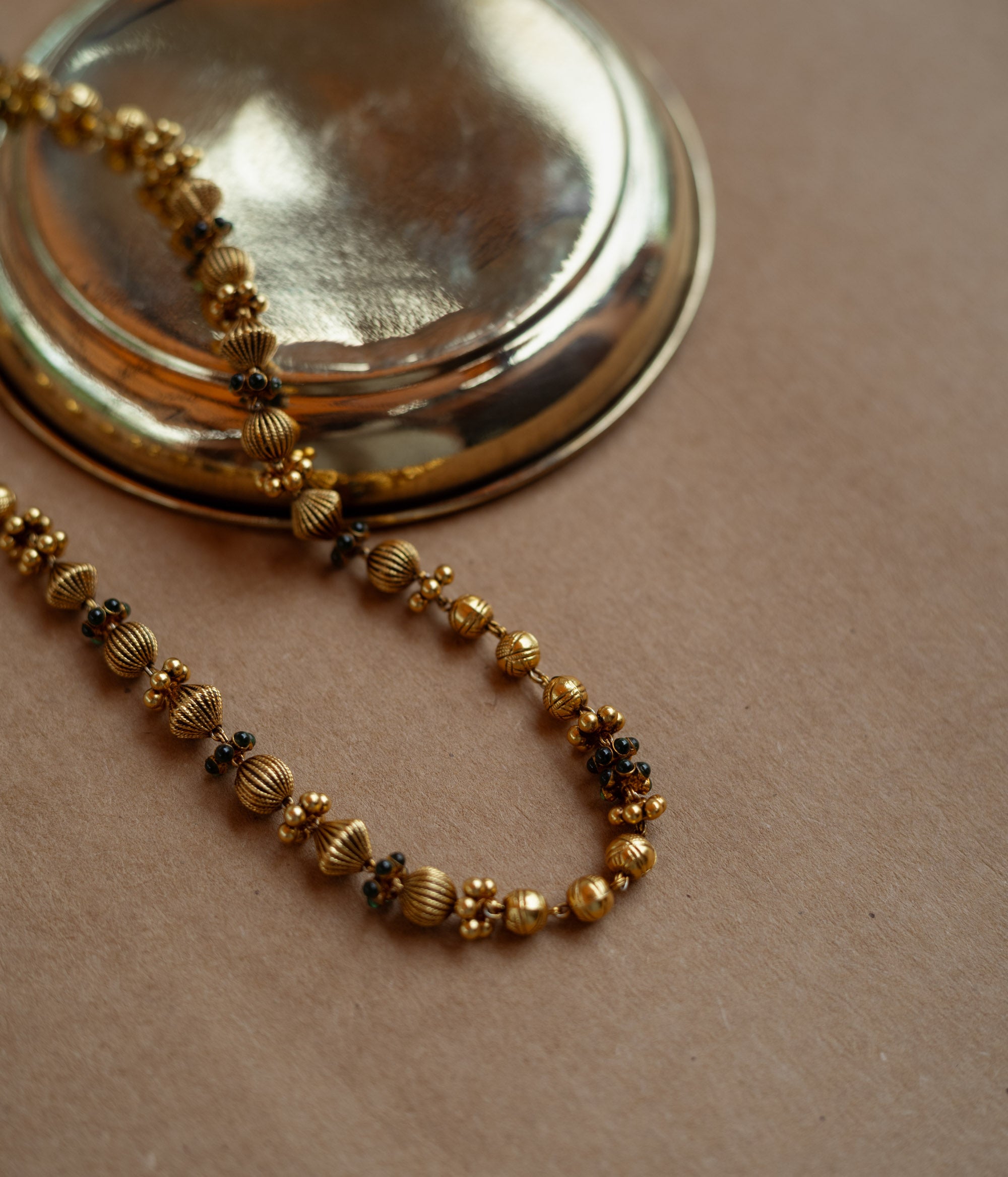 A long, delicate gold chain with various textured beads. Small green stones are interspersed along the chain, which is draped over a golden plate on a brown surface.
