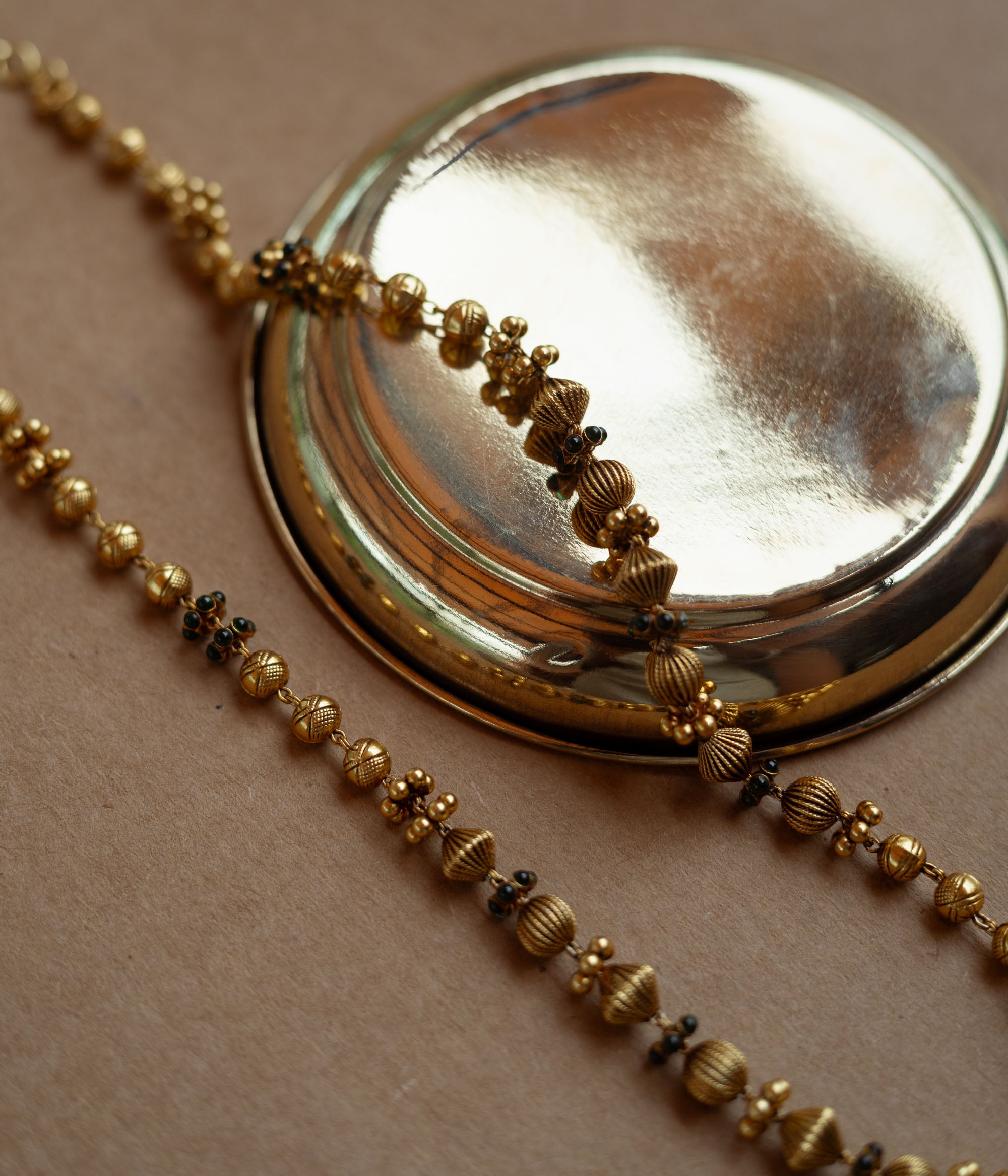 A long, delicate gold chain with various textured beads. Small green stones are interspersed along the chain, which is draped over a golden plate on a brown surface.
