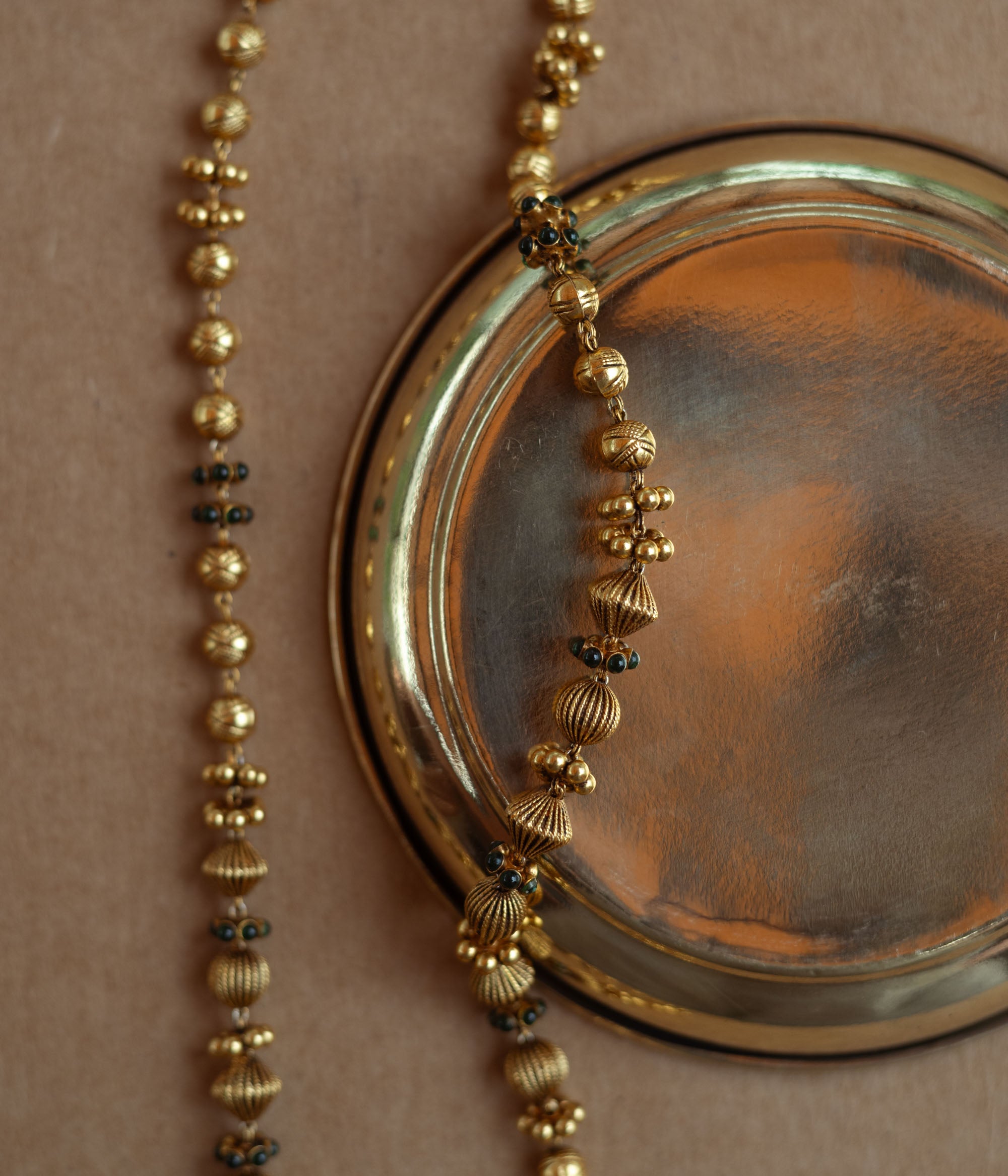 A long, delicate gold chain with various textured beads. Small green stones are interspersed along the chain, which is draped over a golden plate on a brown surface.