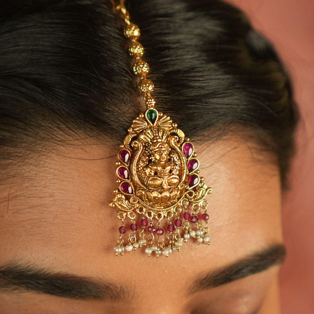 Sacred grace reborn! This gold-plated Lakshmi maangtika, adorned with red and green Kundan stones, glows with white and pink bead droplets—radiating divine blessings and timeless elegance.