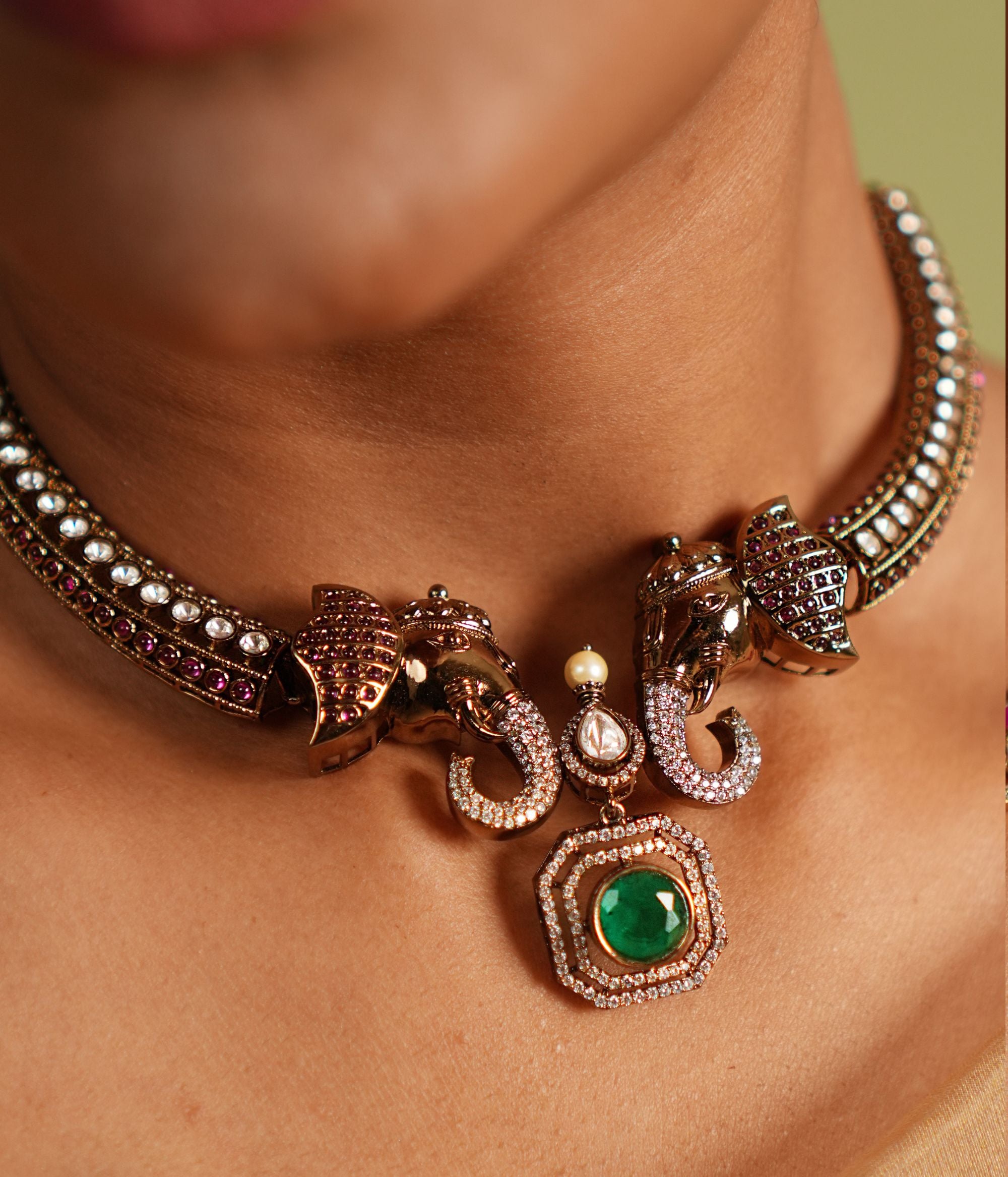 "Gold-plated silver Kanti necklace featuring an intricately designed elephant-shaped pendant. The elephant's ears are adorned with vibrant red cut stones, while its trunk is embellished with shimmering CZ stones. A striking green cut stone drop, encircled by CZ stones, adds an elegant finish to the design, making this necklace a perfect blend of tradition and sophistication.