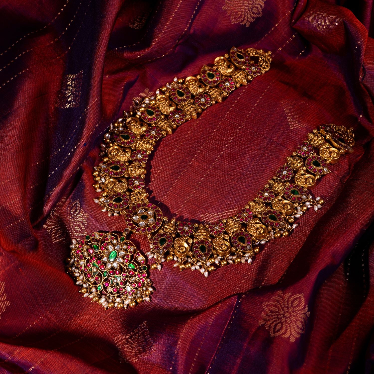 Antique gold-plated silver temple necklace featuring a detailed central pendant with intricate floral designs, inlaid with red, white, and green Kundan stones. The design showcases divine Goddess Lakshmi and elegant peacock motifs, complemented by smaller hanging pendants along the lower edge. Adorned with delicate white pearls and golden beads, this masterpiece exudes traditional charm and regal elegance.