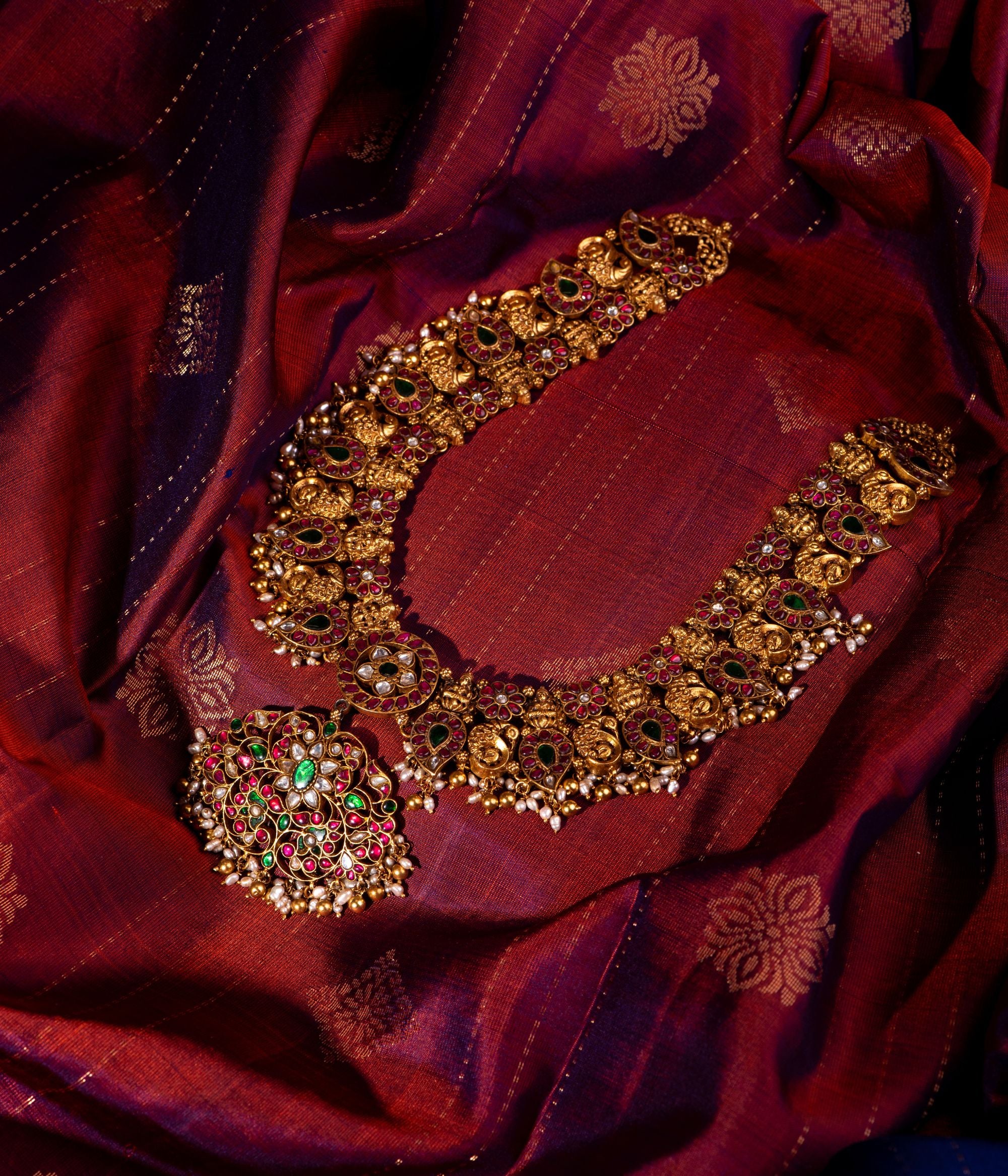 Antique gold-plated silver temple necklace featuring a detailed central pendant with intricate floral designs, inlaid with red, white, and green Kundan stones. The design showcases divine Goddess Lakshmi and elegant peacock motifs, complemented by smaller hanging pendants along the lower edge. Adorned with delicate white pearls and golden beads, this masterpiece exudes traditional charm and regal elegance.